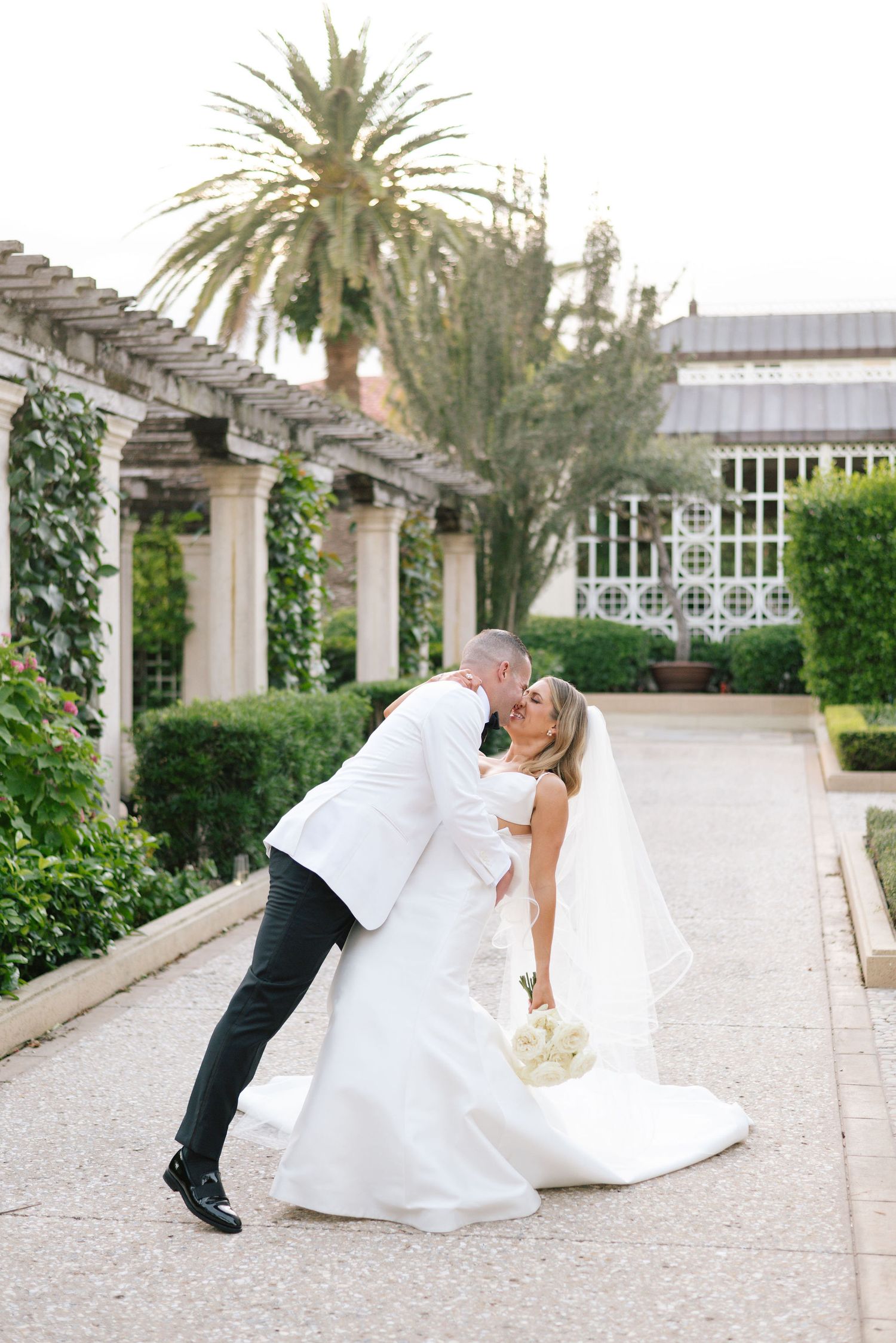 A couple shares a romantic kiss under a pergola with palm trees and greenery in an elegant garden setting.