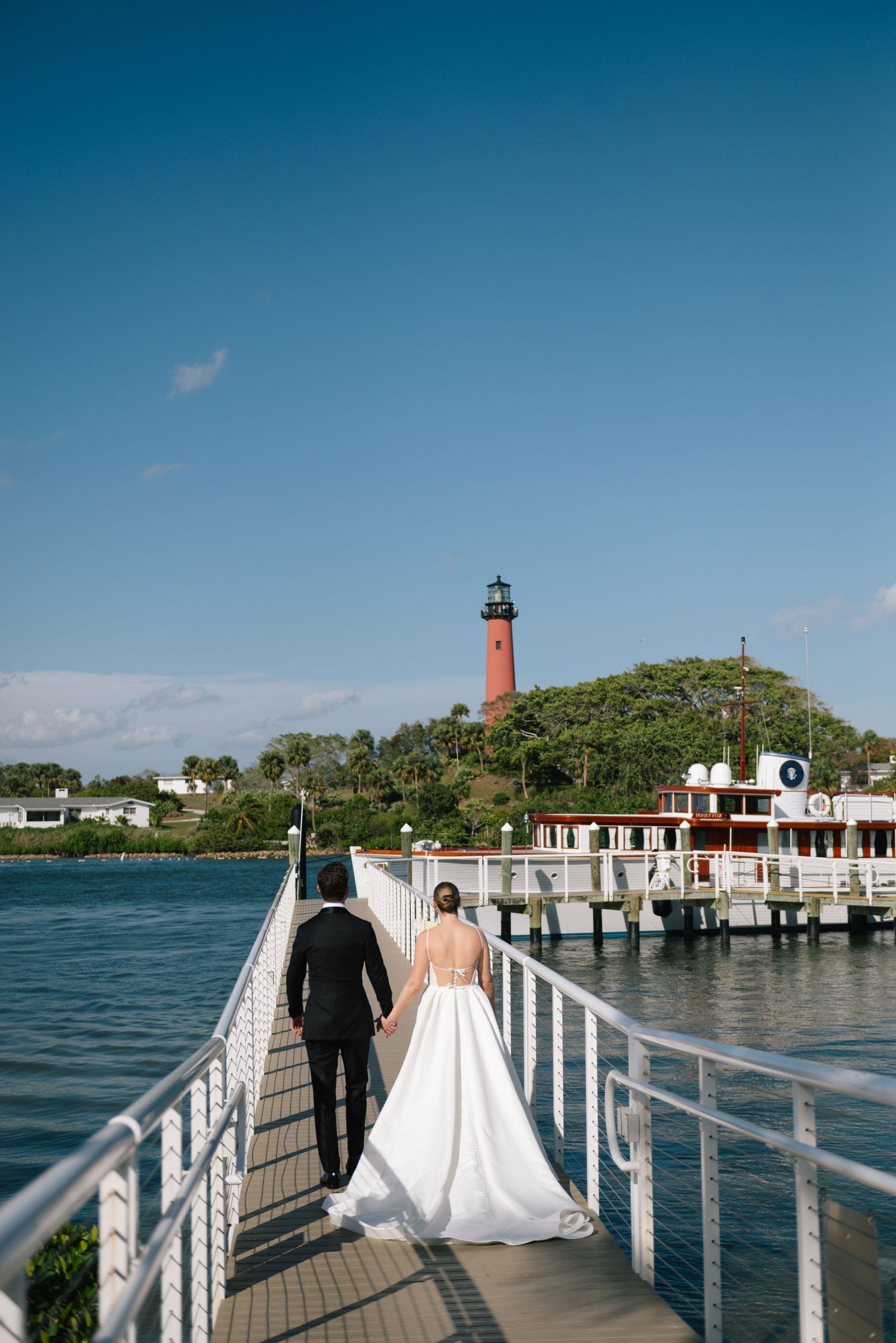 Wedding couple walking on waterfront dock with lighthouse and coastal buildings in background.