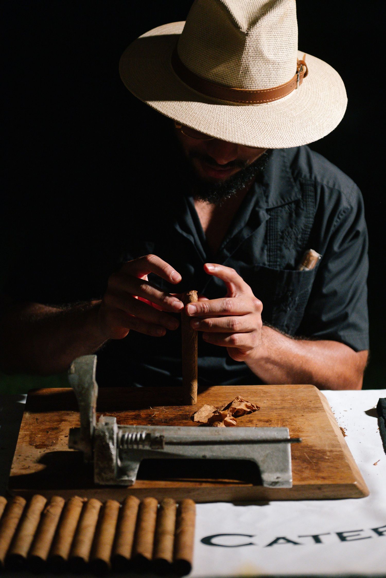 Artisan in panama hat hand-rolling cigars at wooden table during wedding reception.