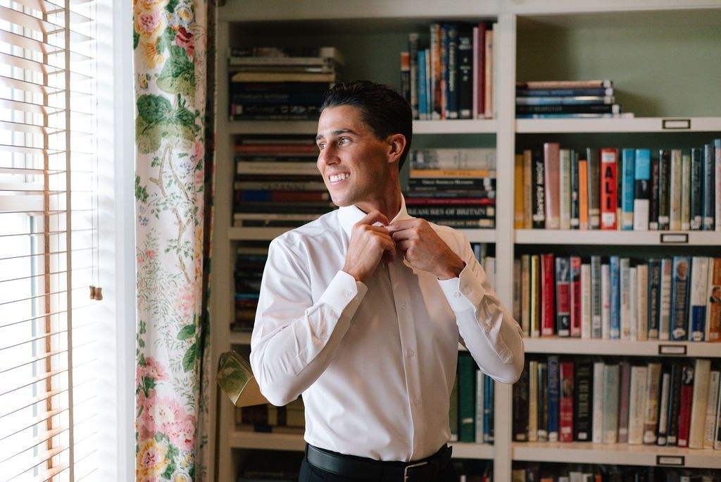 Person in white shirt adjusting collar by window with bookshelf background.