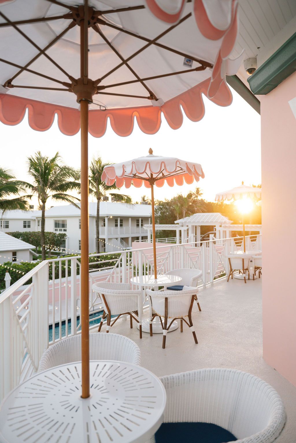 Coral-trimmed umbrellas shade white outdoor furniture on a seaside terrace at sunset.