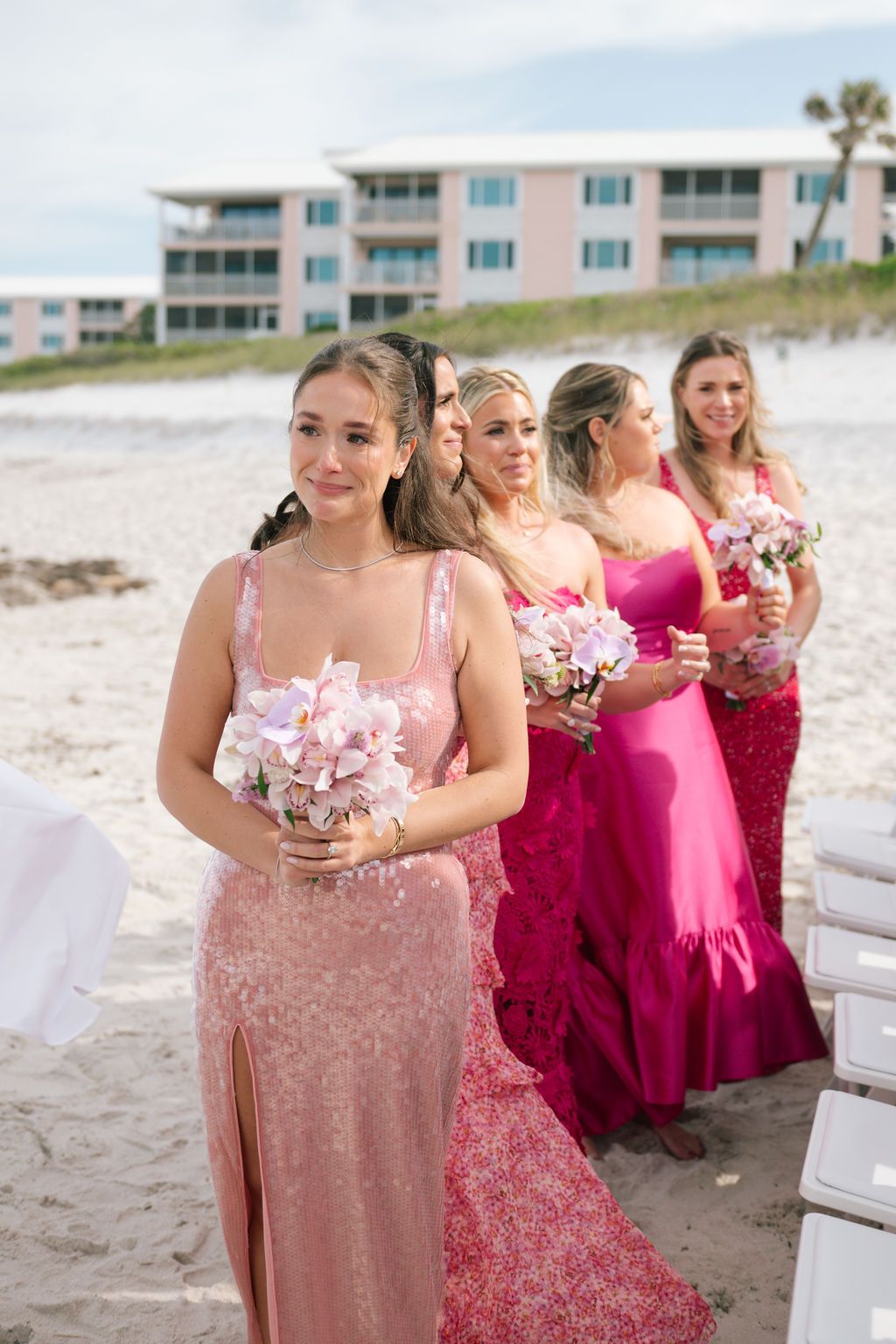 Bridesmaids in pink and burgundy dresses walking on beach with drinks.