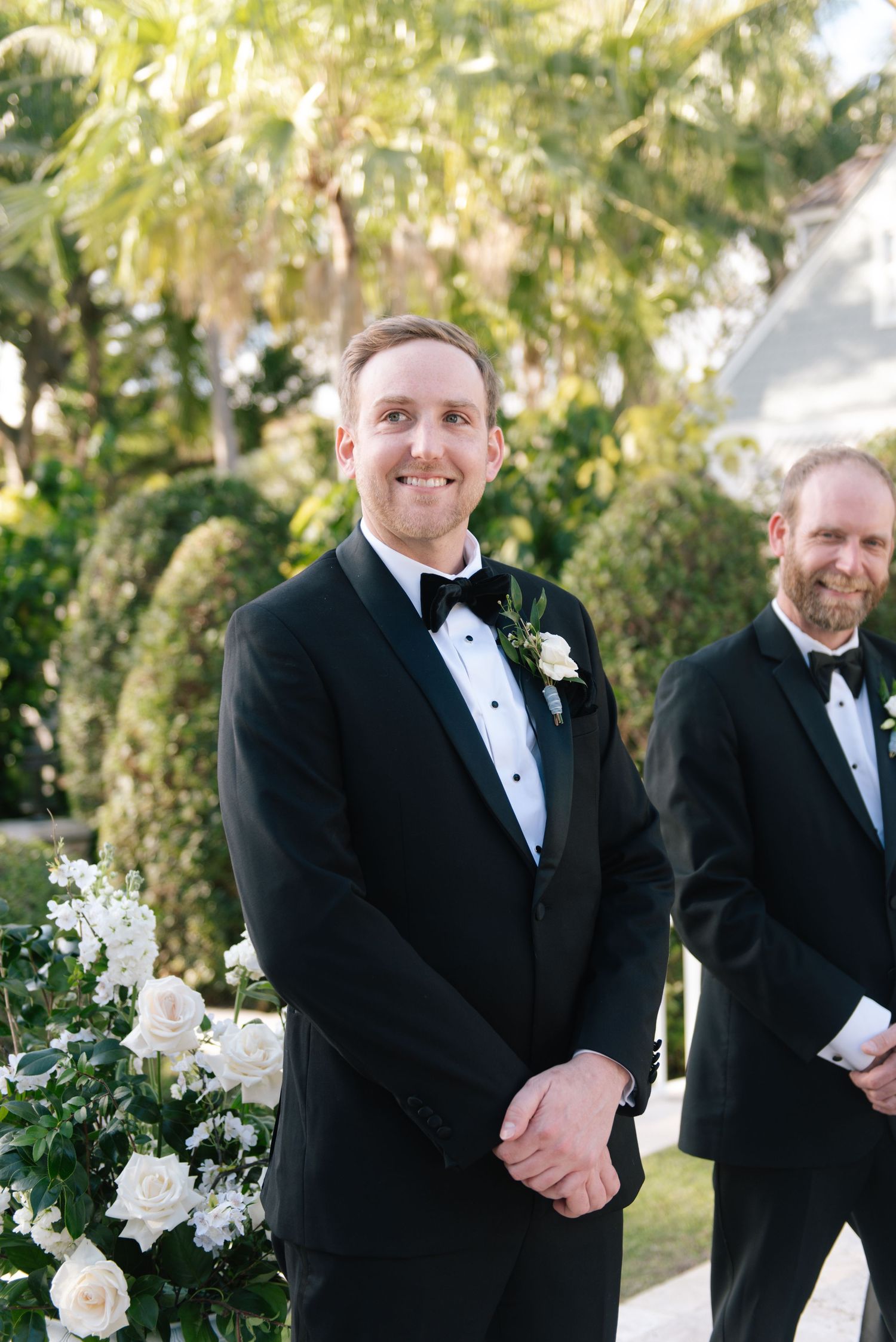 Groomsmen in black tuxedos stand together at an outdoor wedding ceremony.