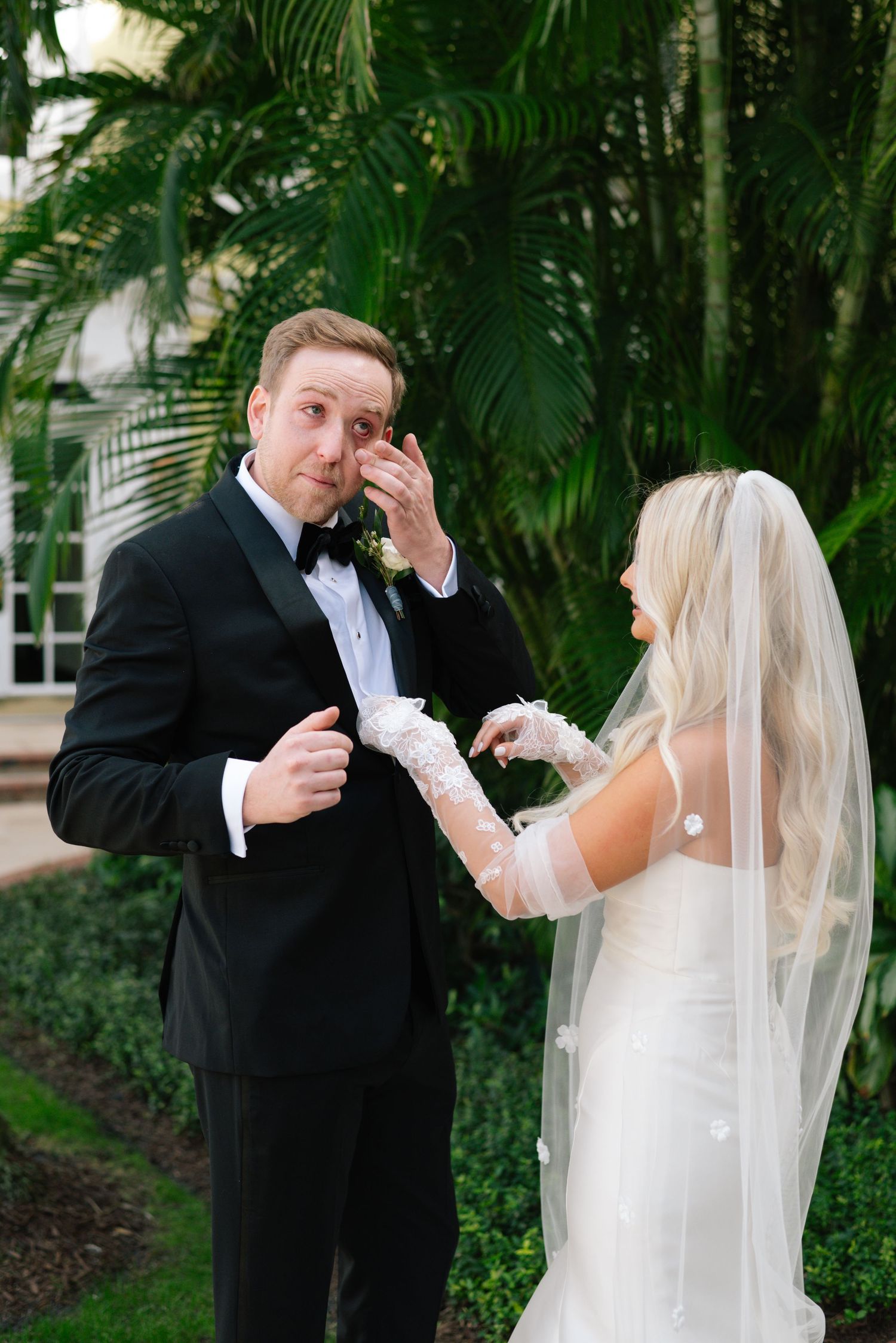 Bride and groom share an emotional first look moment surrounded by tropical palm trees in a garden setting.