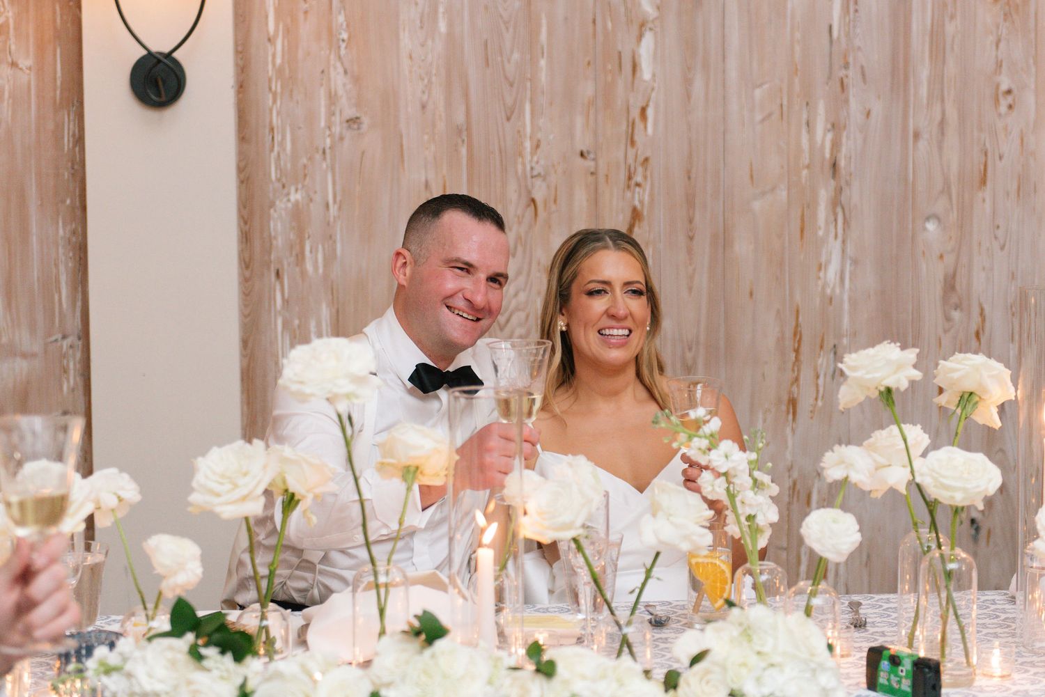 A couple sits at their wedding reception table decorated with delicate white flowers and elegant place settings.