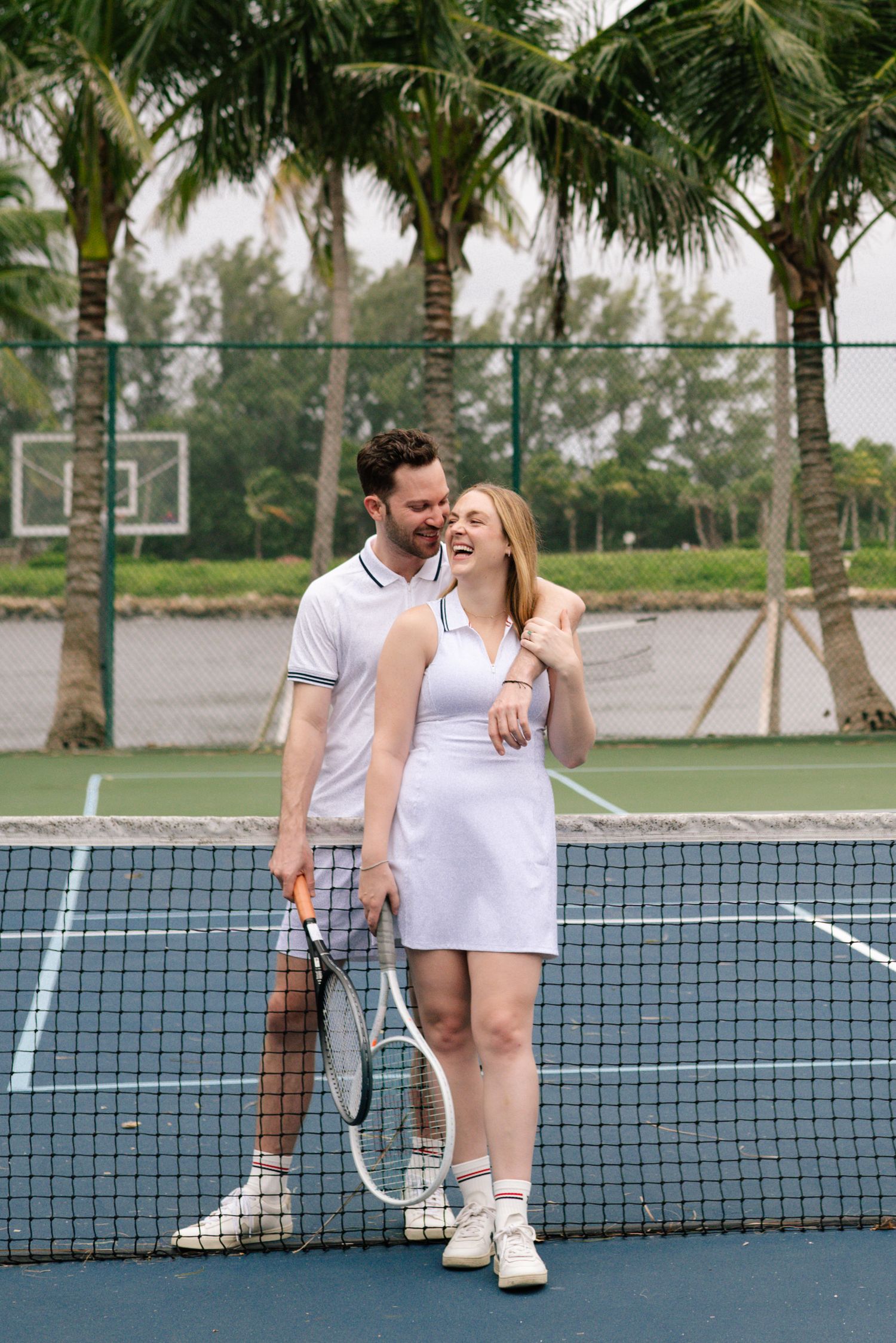 A couple in white tennis outfits share romantic moments on a palm tree-lined tennis court.
