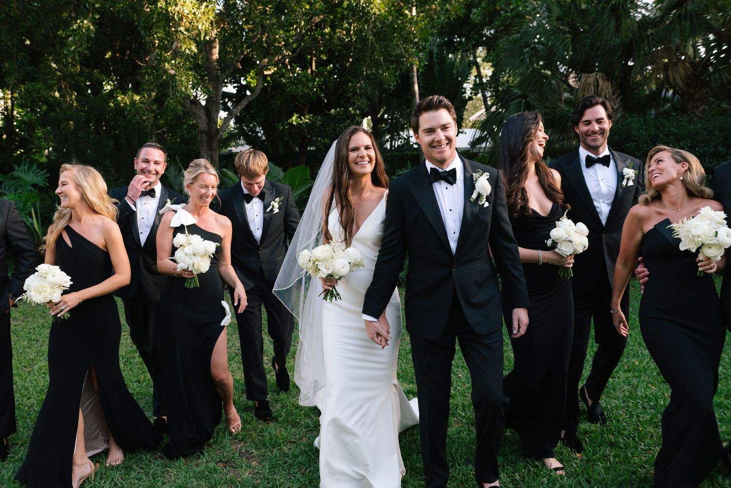 Wedding party in formal black and white attire posing together in a garden setting.