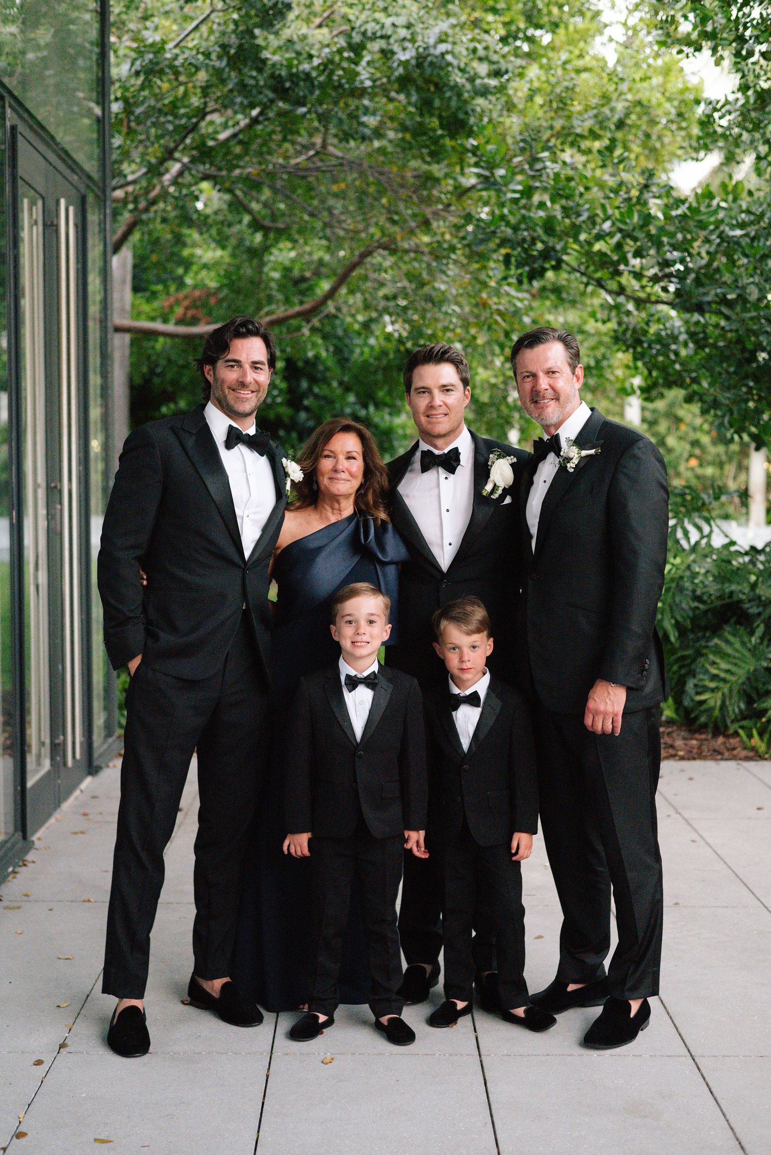 Wedding party in formal black tuxedos and evening wear pose together for a group photo outdoors.