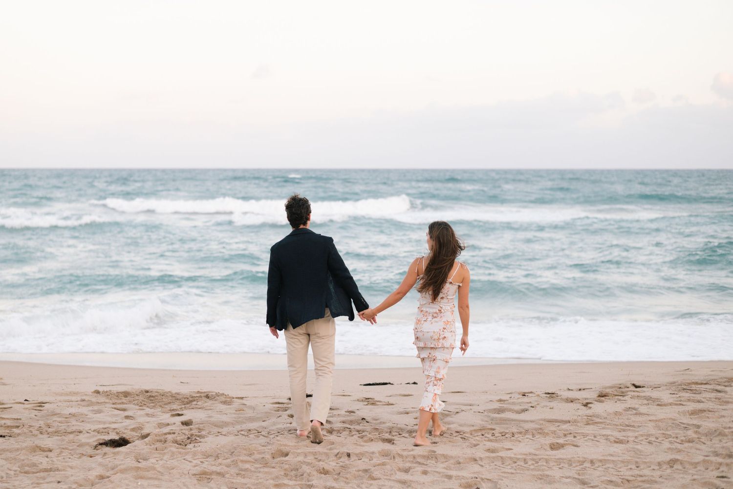 A couple walks hand in hand along a serene beach with ocean waves crashing in the background.