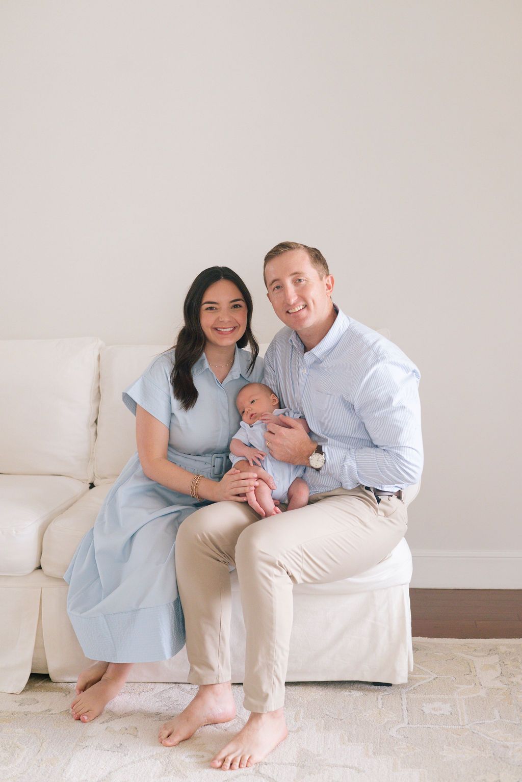 Family portrait on white couch showing parents with their newborn in coordinating blue and white outfits.