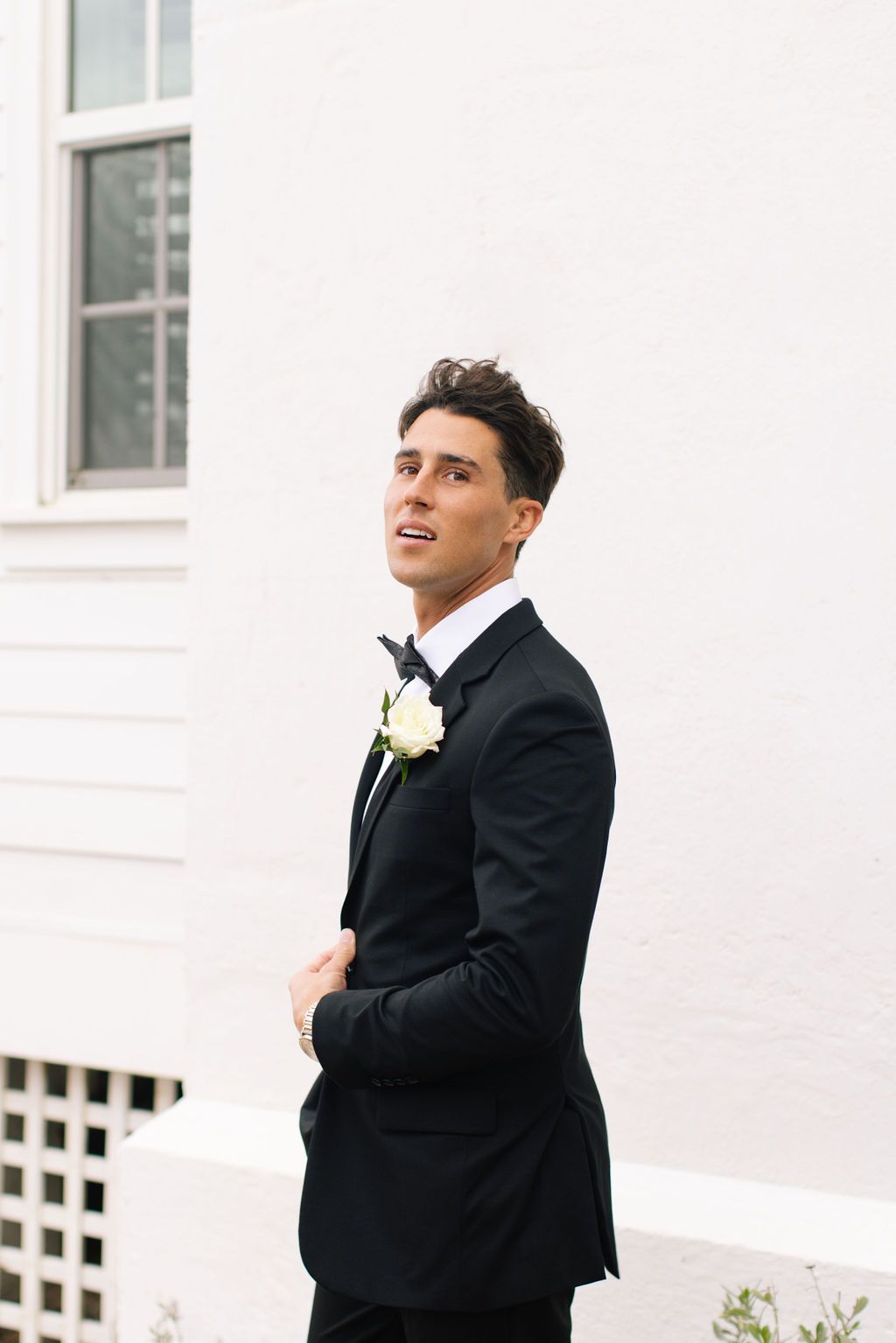 Groom in classic black tuxedo poses joyfully against white building backdrop.