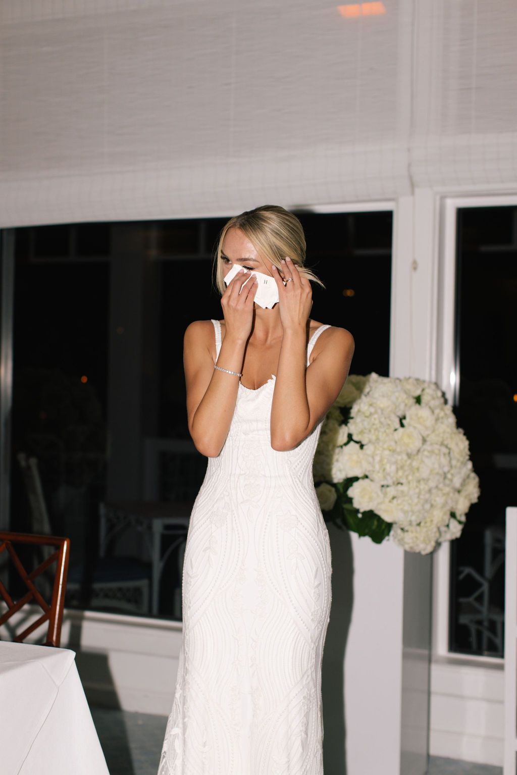Emotional bride in fitted white dress wipes tears with tissue during wedding reception.
