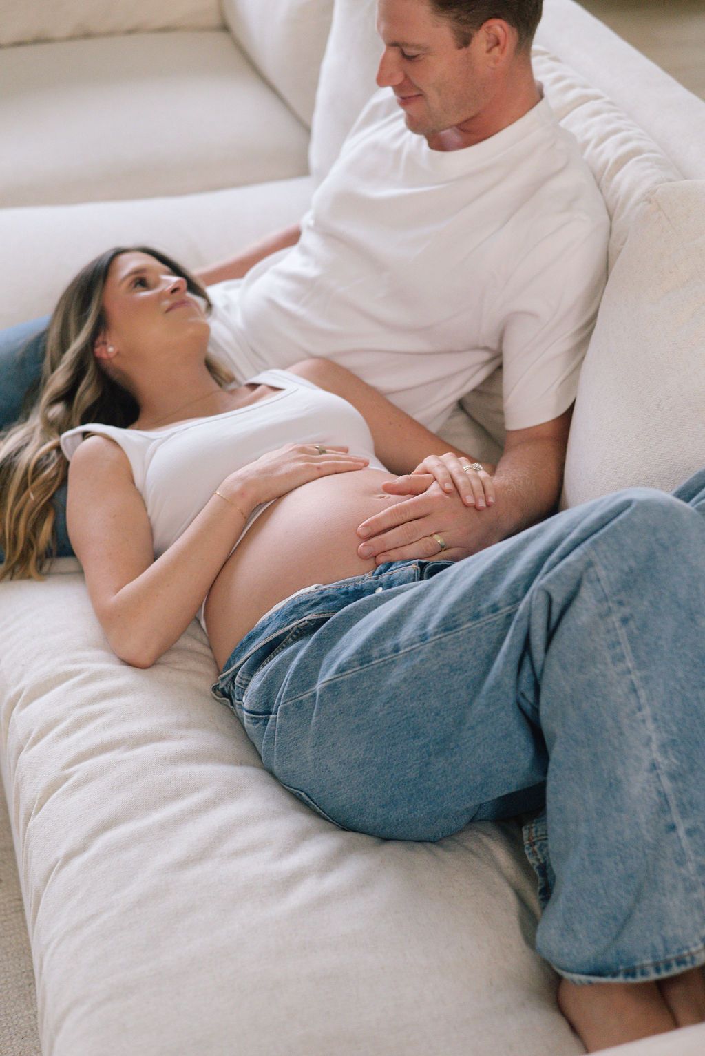 Expecting couple relaxing together on white sofa during pregnancy photoshoot.