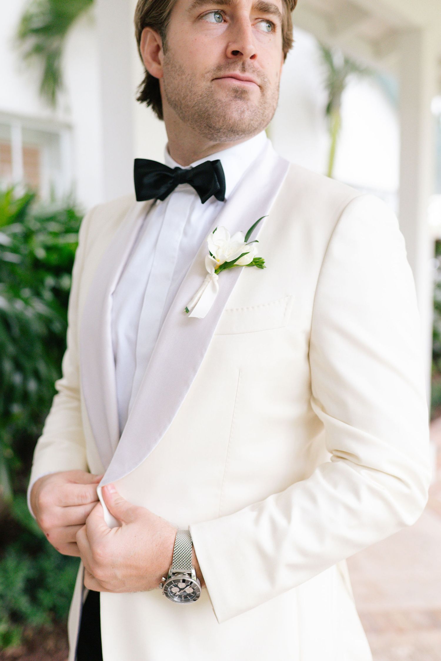 Groom in white dinner jacket with black bow tie and boutonniere poses in elegant setting.