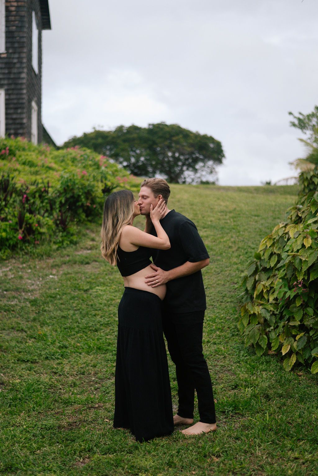 Couple embraces on a grassy slope with trees and foliage in the background.