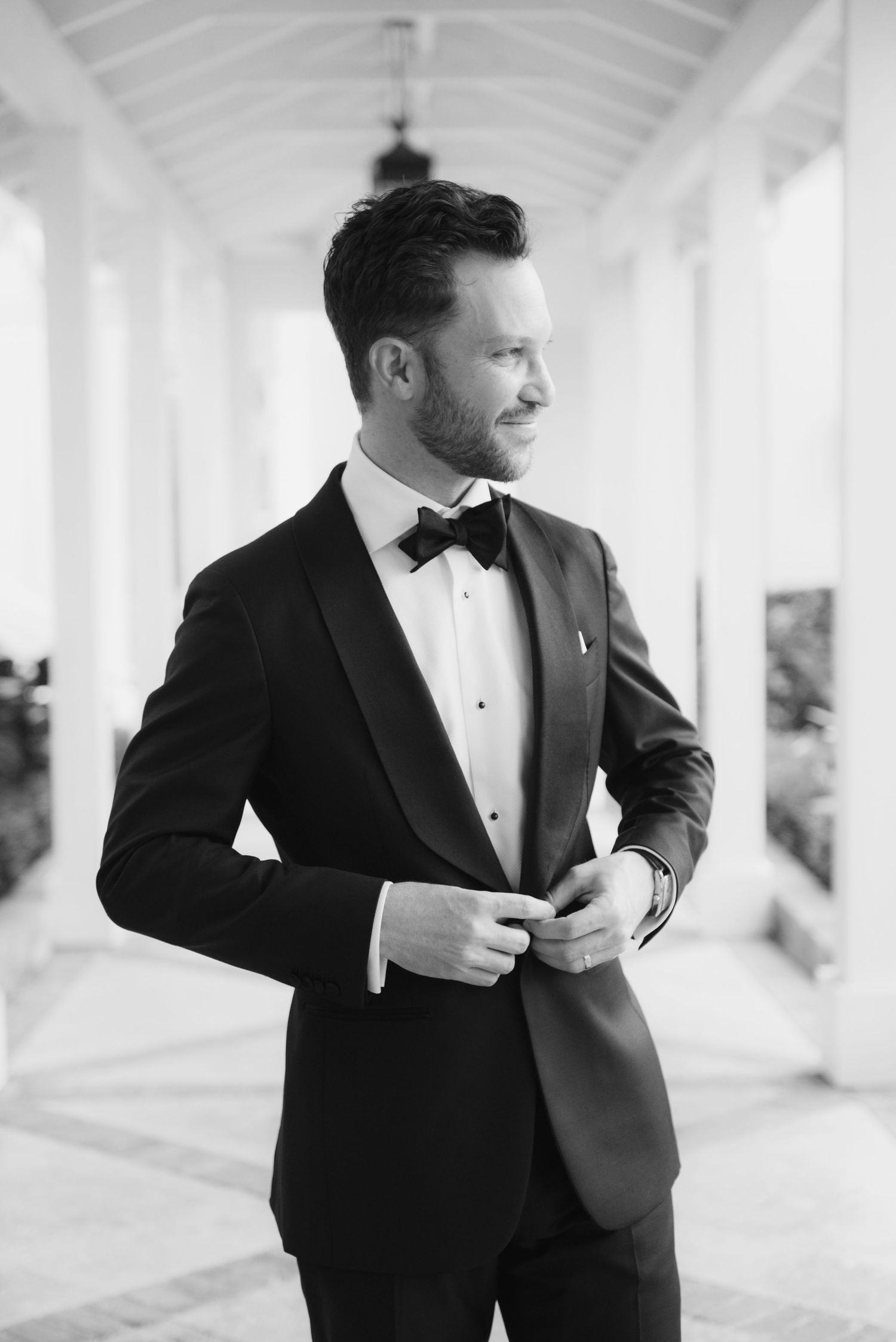 Classic black and white portrait of a groom in a tuxedo and bowtie standing in an elegant hallway.