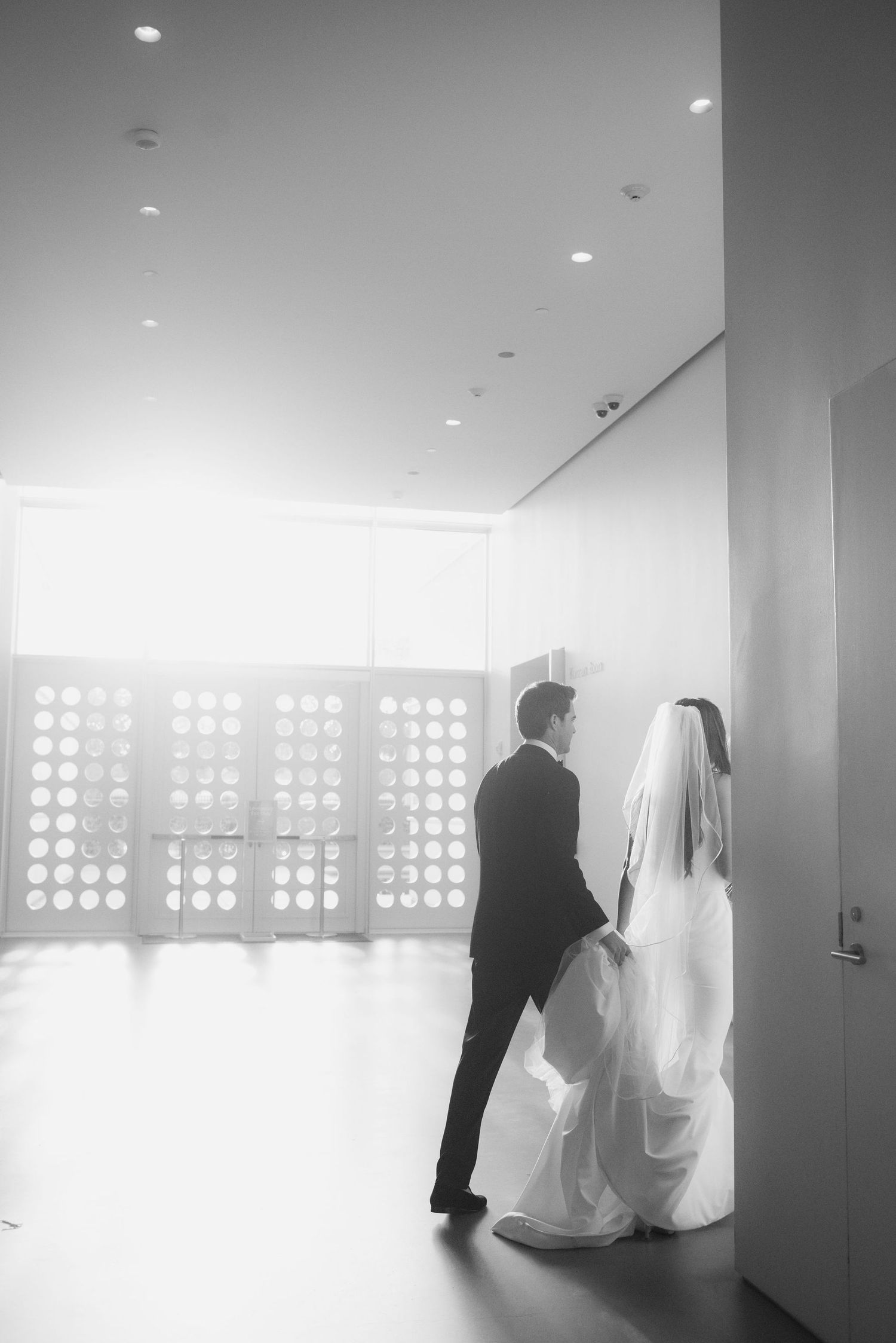 A minimalist wedding scene in a modern white hallway with circular window patterns creating dramatic lighting.