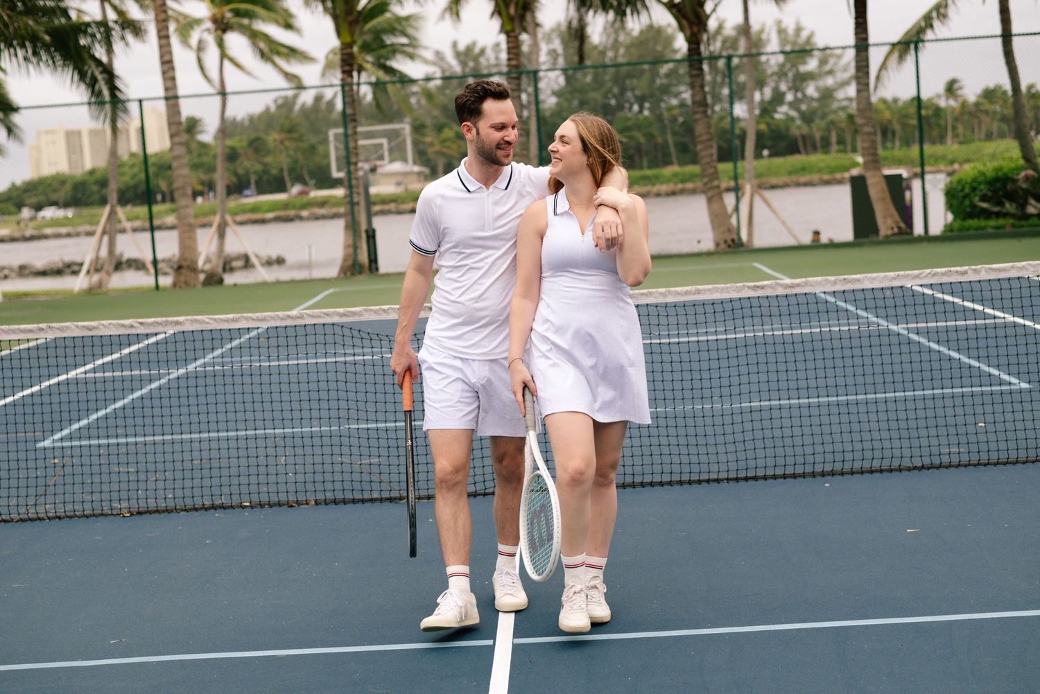 A couple in white tennis outfits walking together on an outdoor tennis court with palm trees in the background.