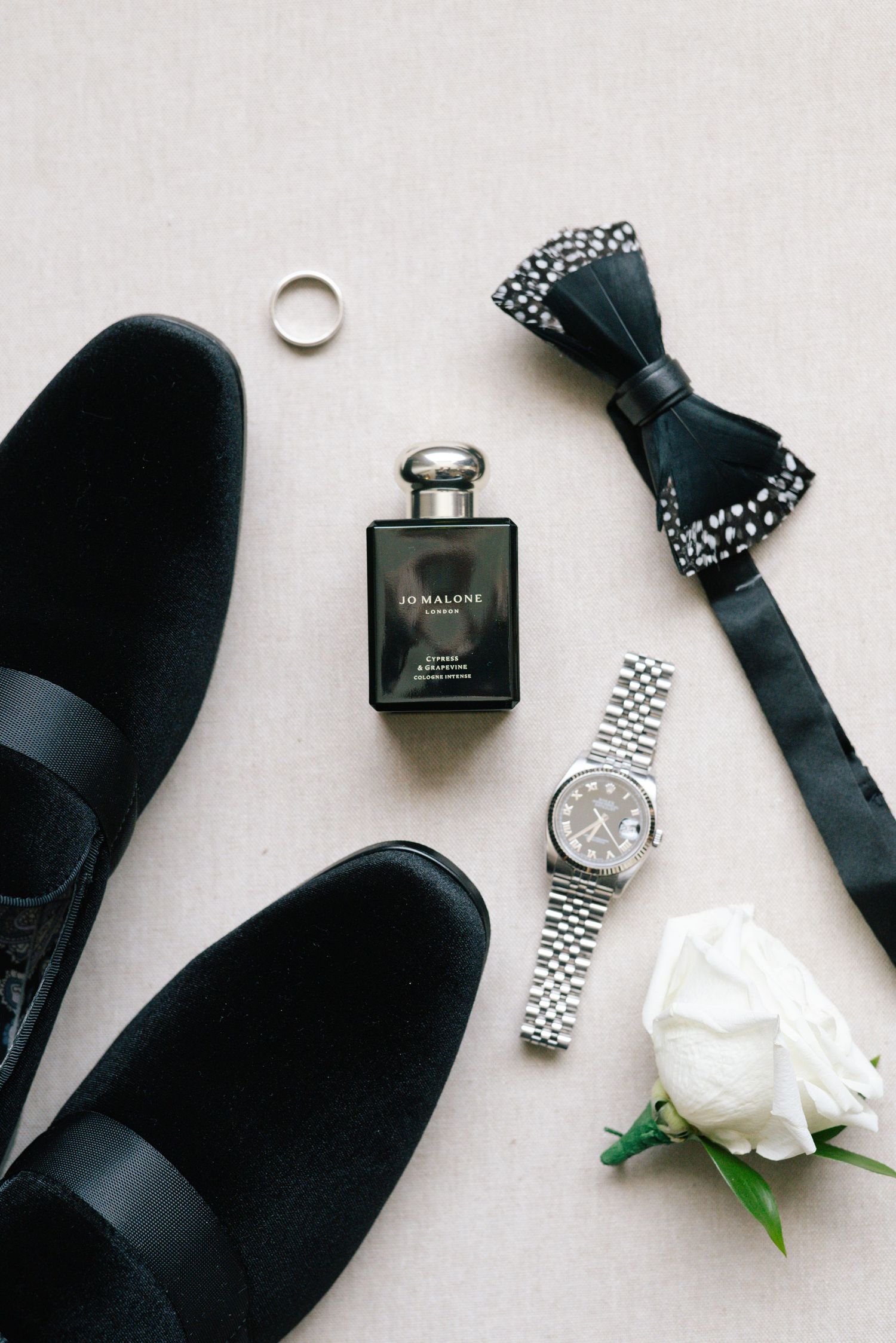Groom wedding flat lay with black loafers, Jo Malone cologne, Rolex watch, bow tie, and rose.