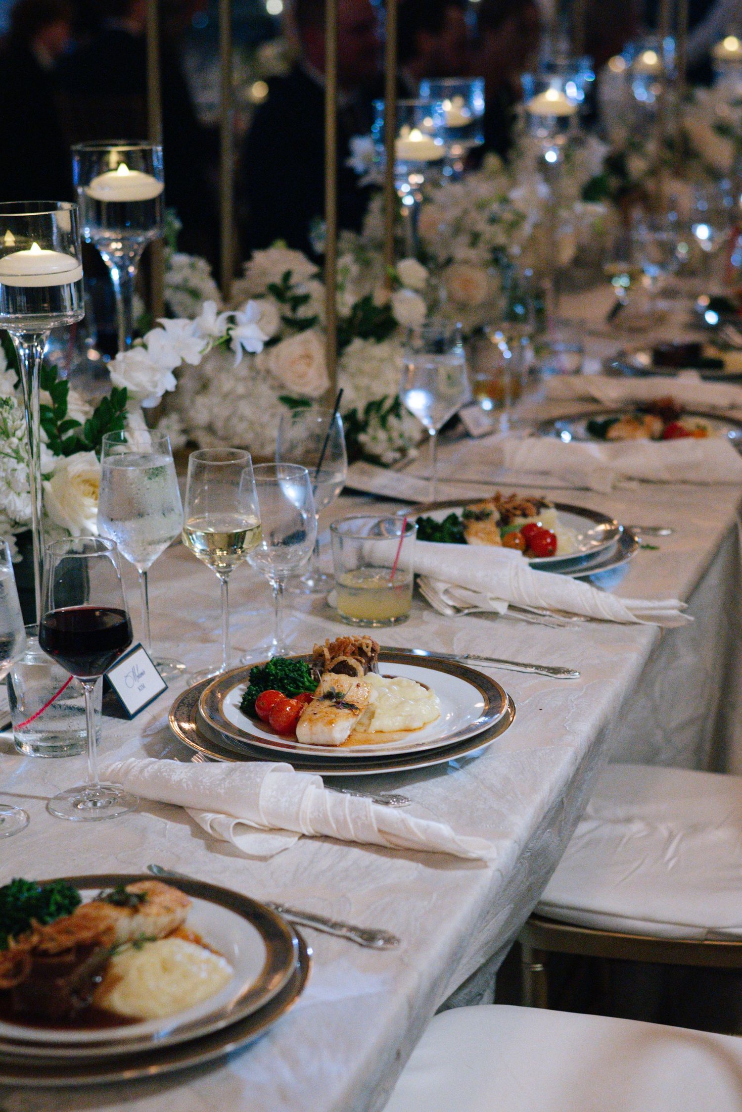 Elegant wedding dinner service with silver chargers, white florals, candles, and plated entrees.
