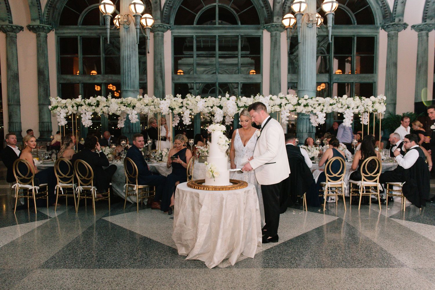 Newlyweds cut wedding cake on dance floor in stunning ballroom with white floral centerpieces.