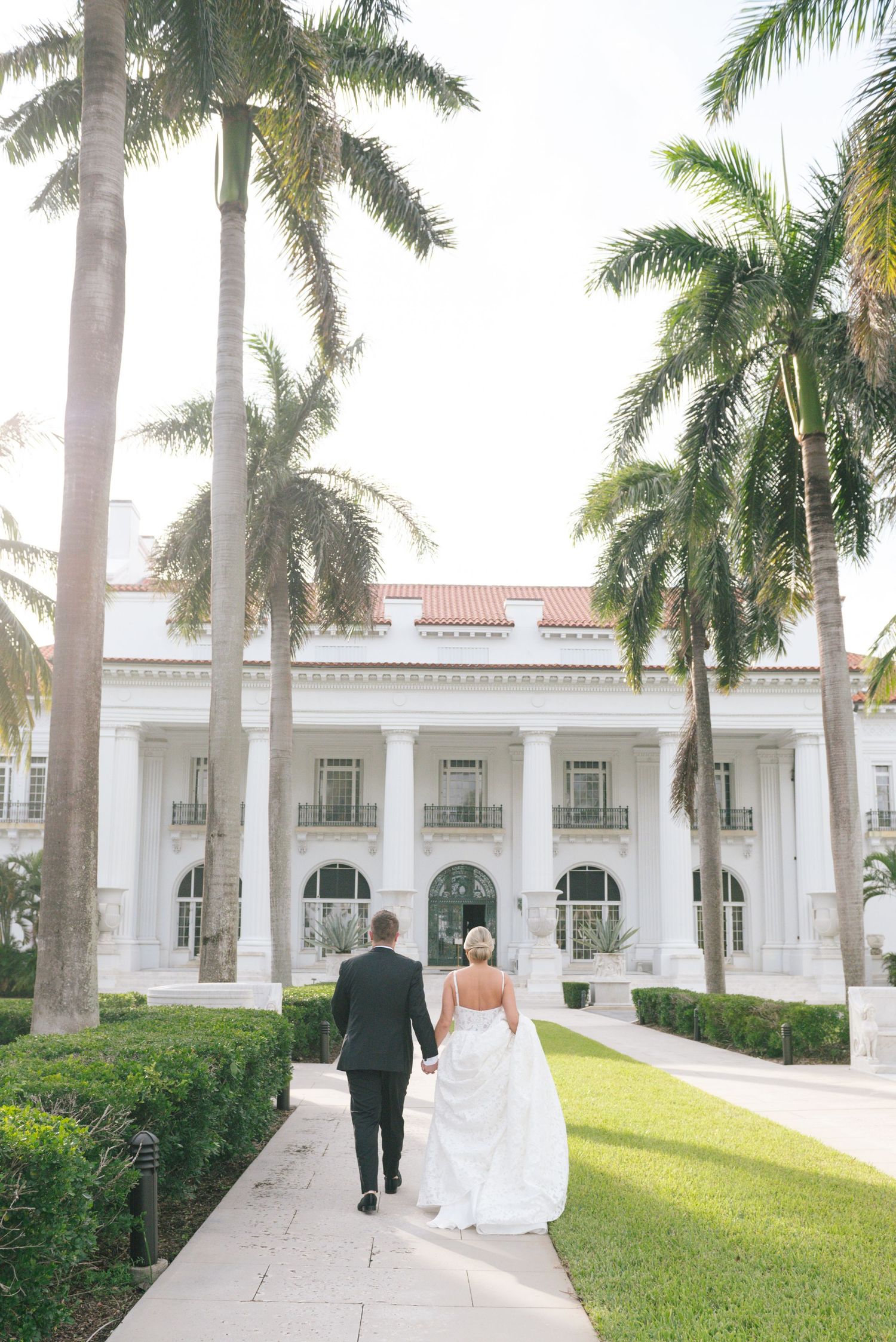 Bride and groom walk hand-in-hand toward a grand white colonial venue lined with tall palm trees on their wedding day.