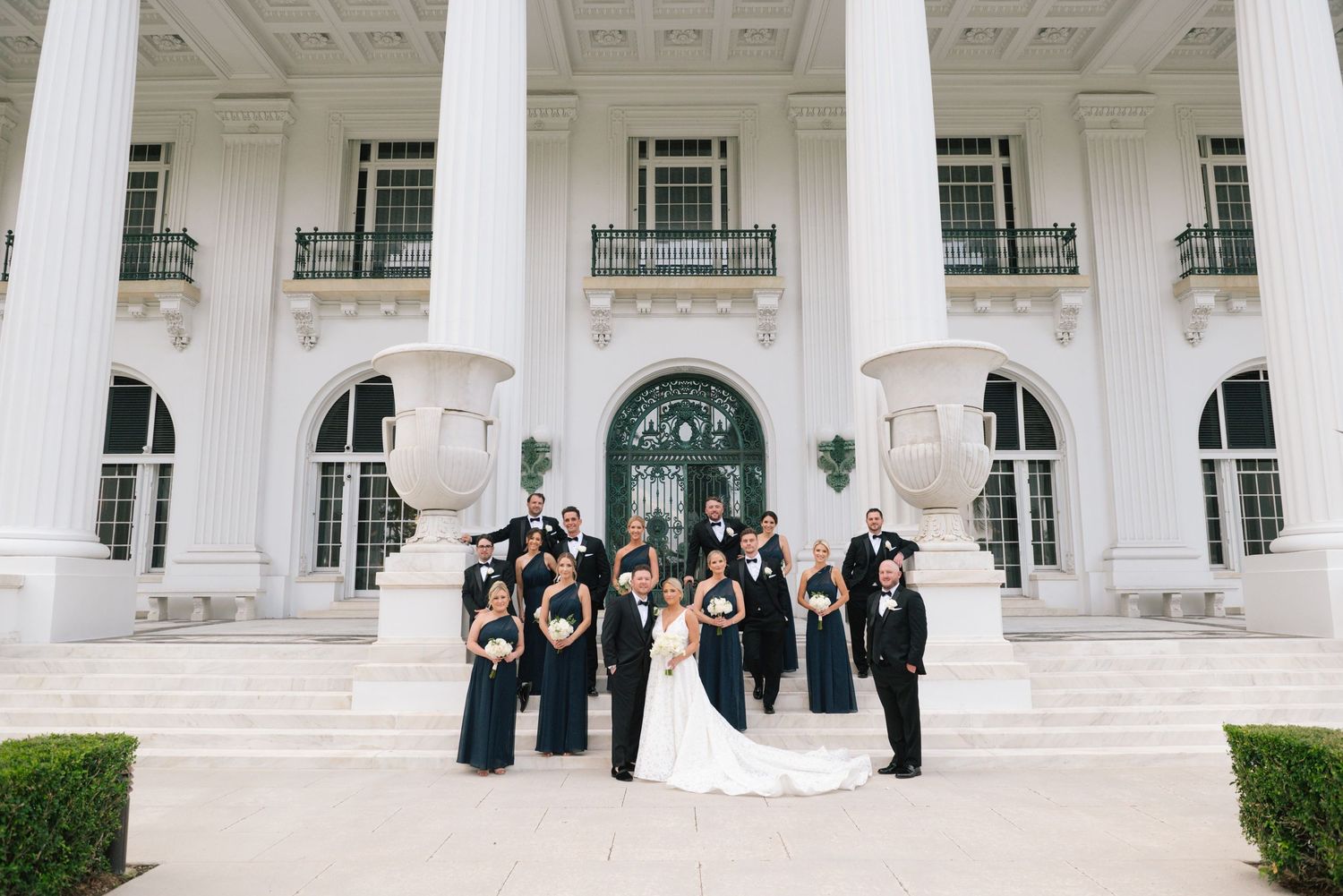 Wedding party in black attire poses on white mansion steps with tall columns.