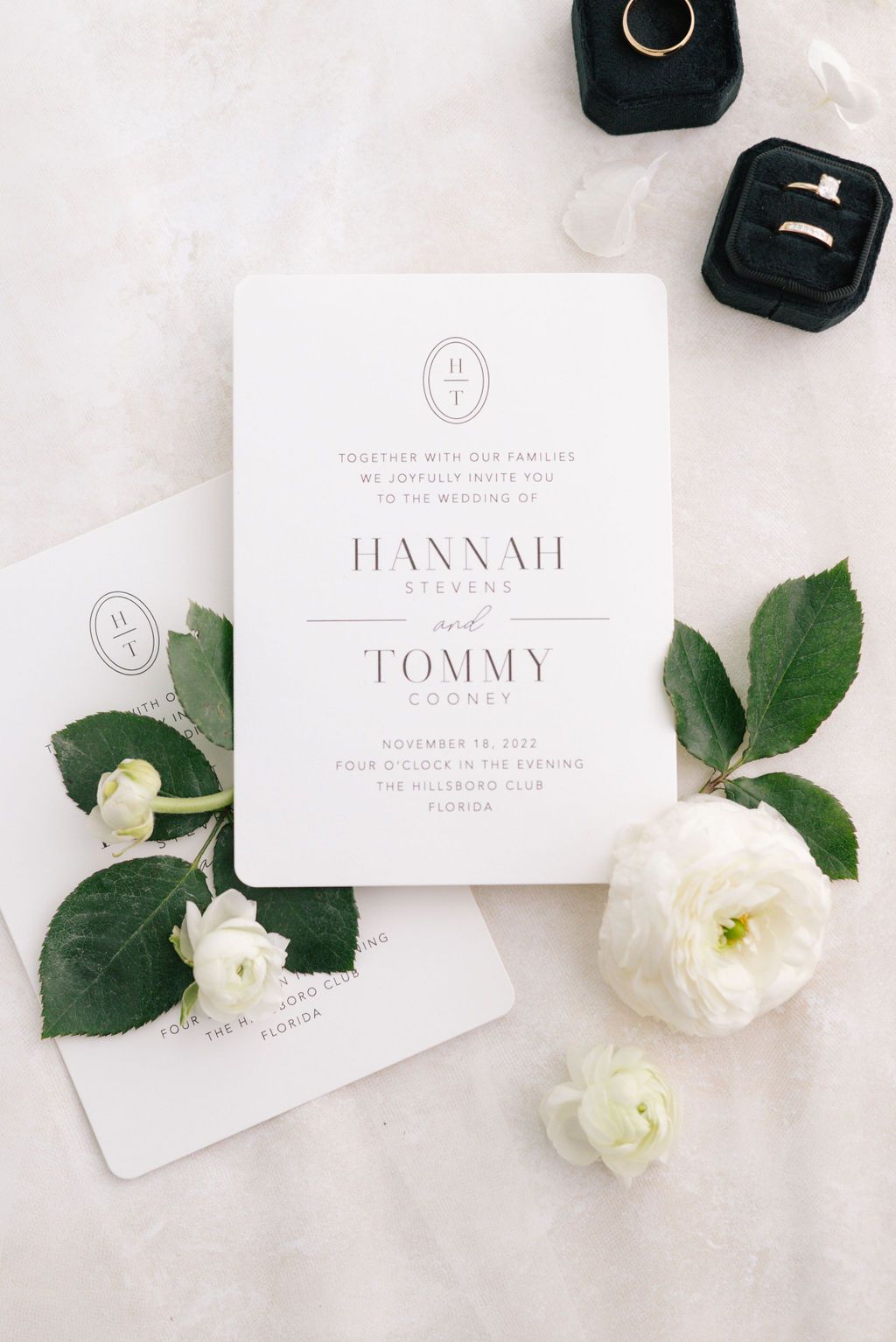 Wedding stationery flat lay with white roses, rings, and black velvet ring boxes.