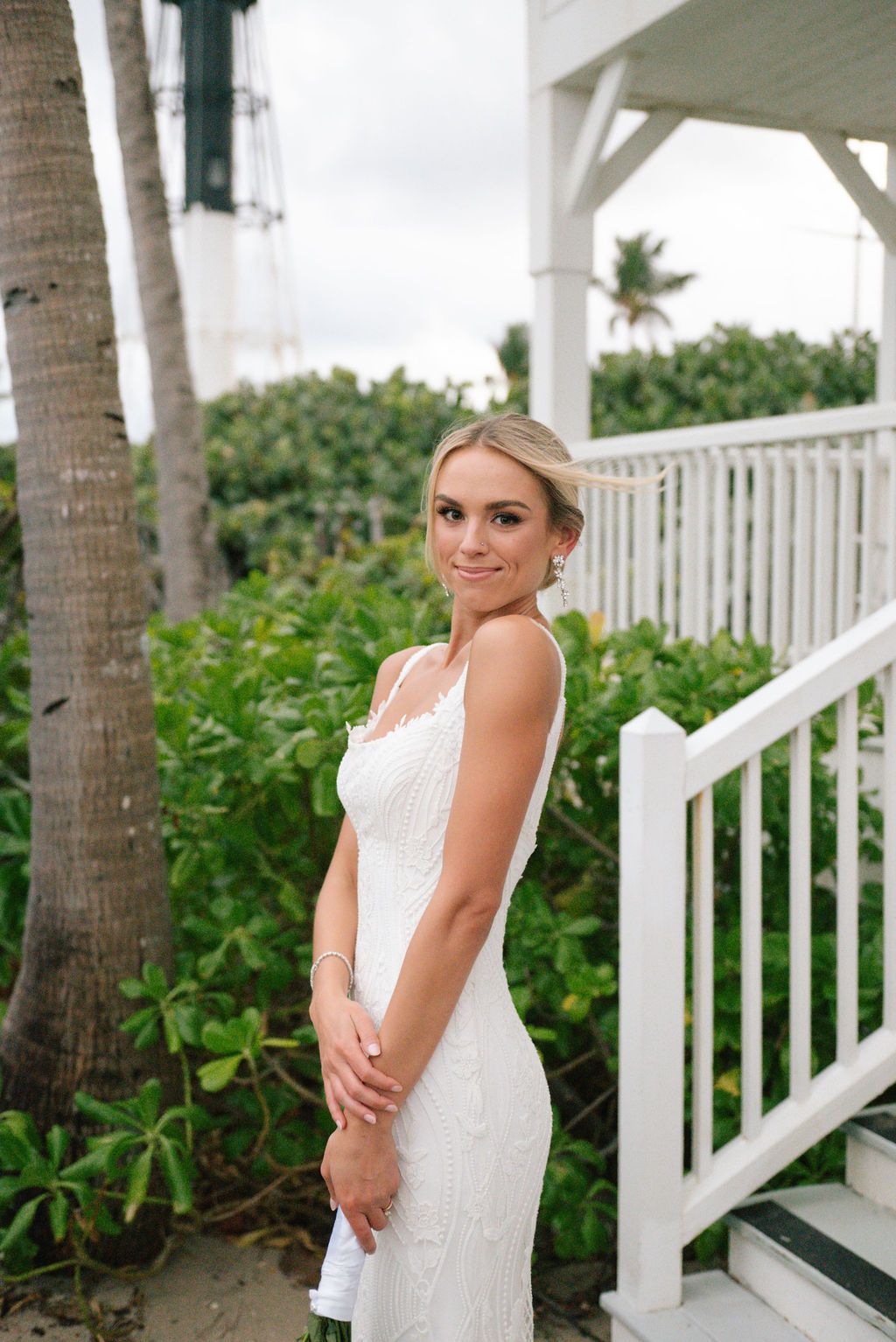 Bride poses in elegant white dress on beachfront porch with tropical landscaping.