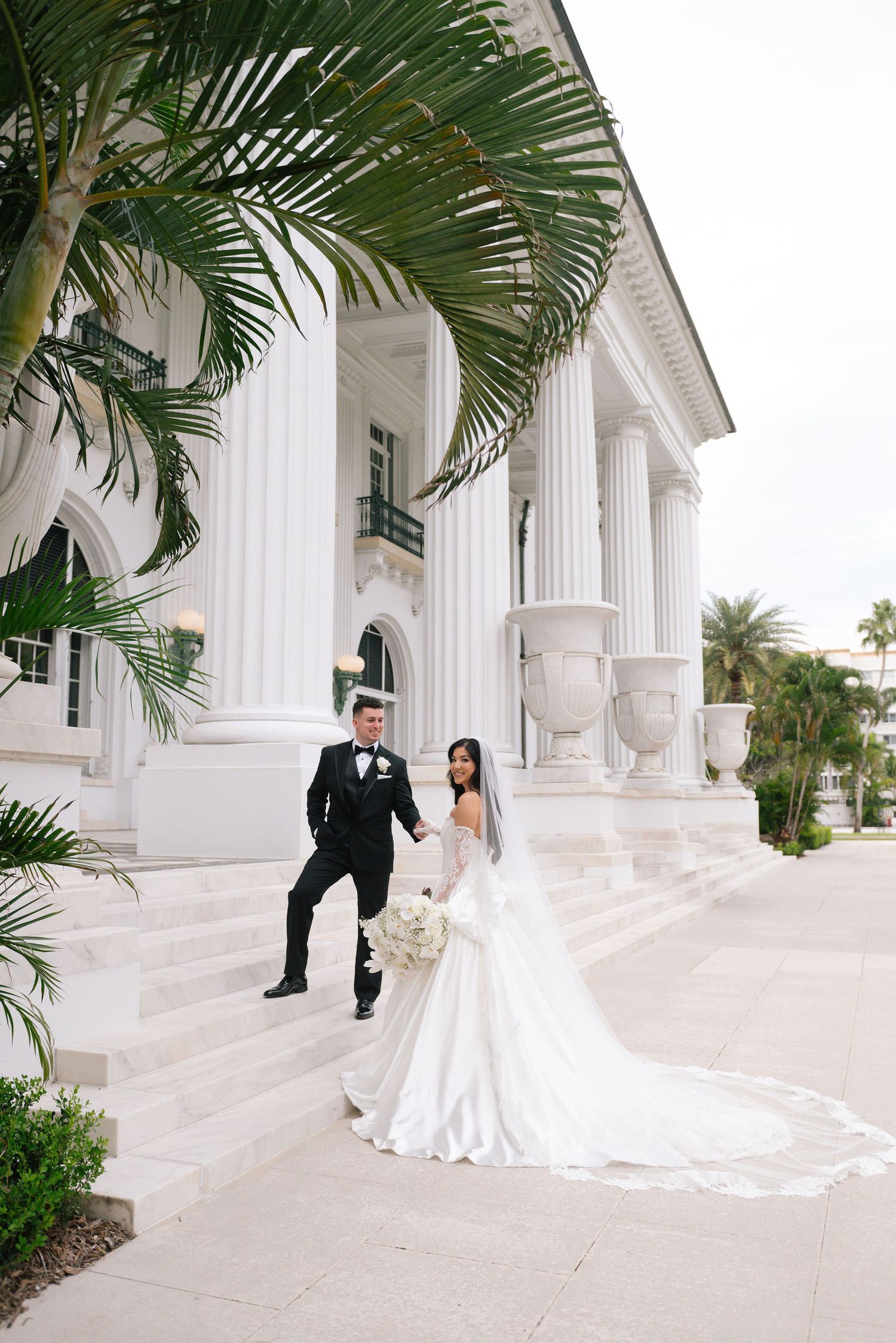 A wedding couple poses between classic white columns with tropical palm fronds overhead.
