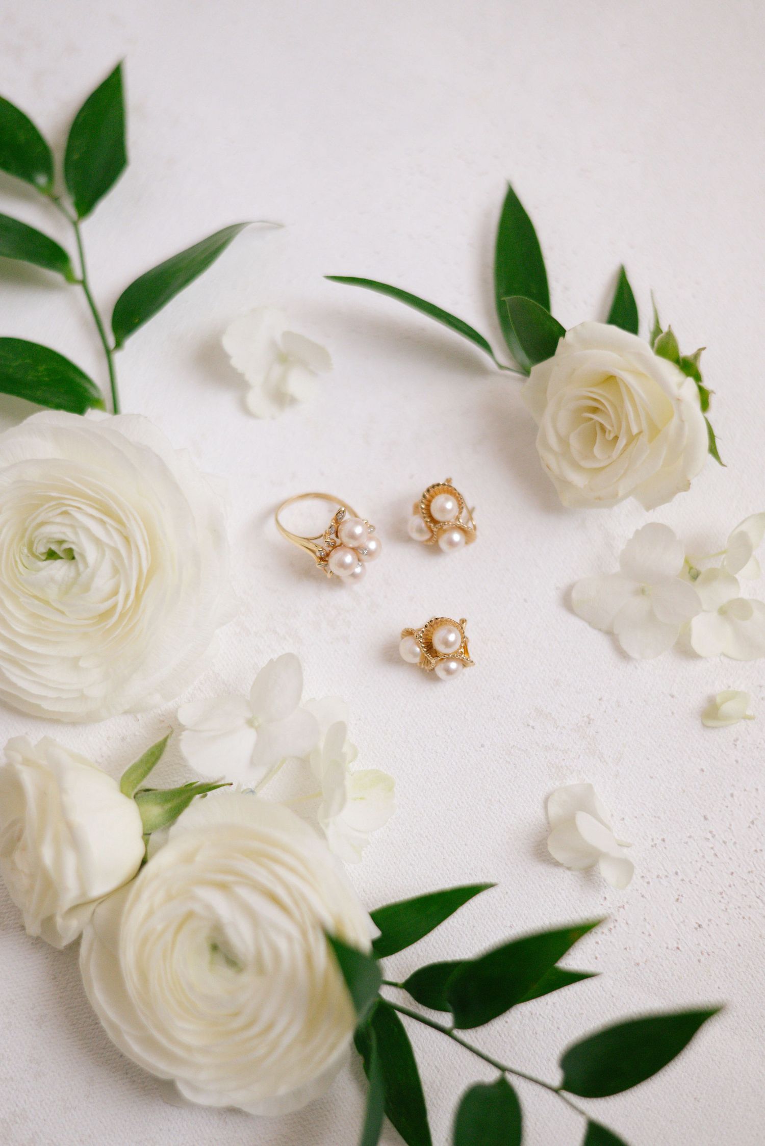 Delicate pearl jewelry set arranged with white ranunculus flowers and green leaves on light background.