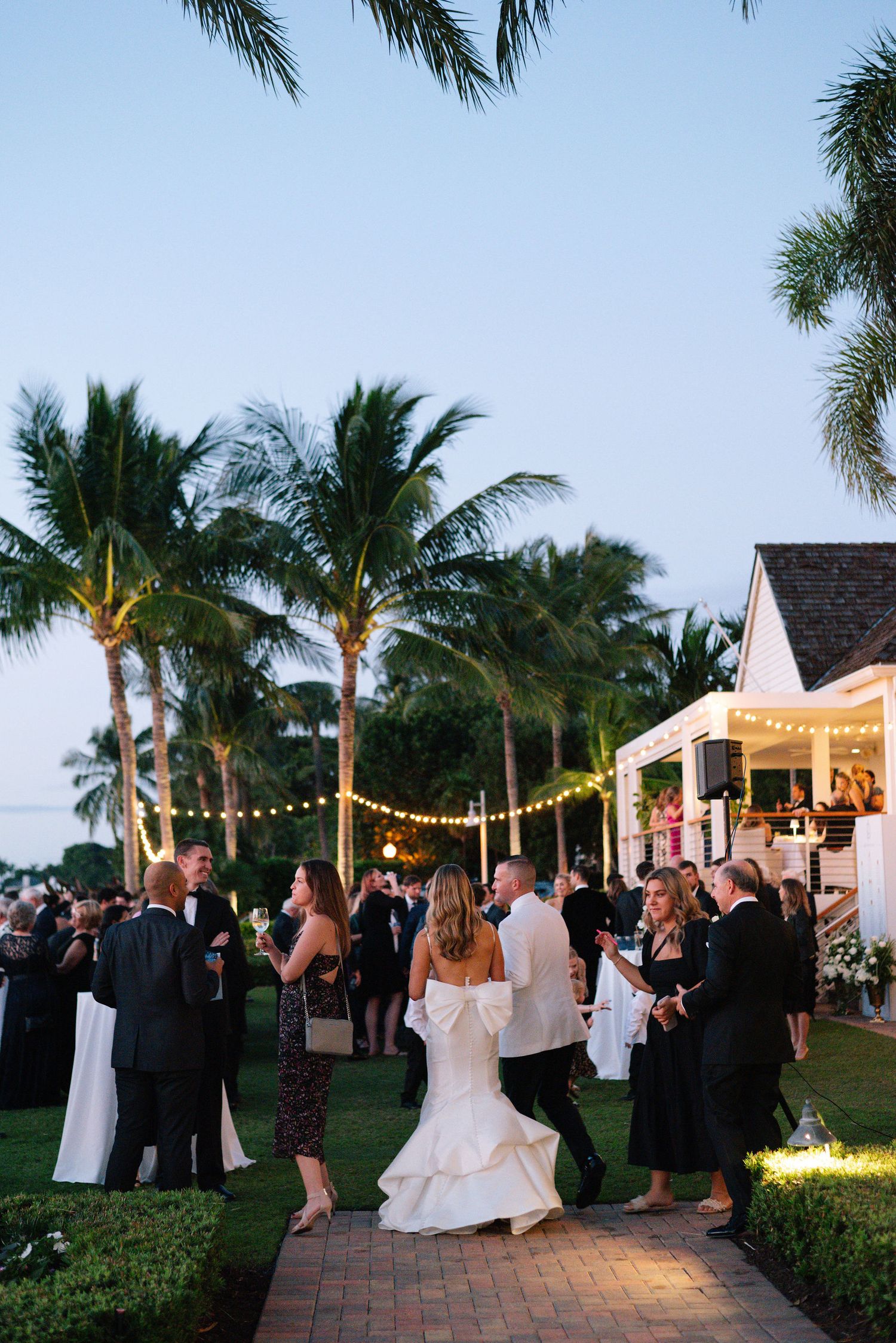 Wedding guests dance and celebrate on an outdoor patio decorated with string lights and palm trees at dusk.