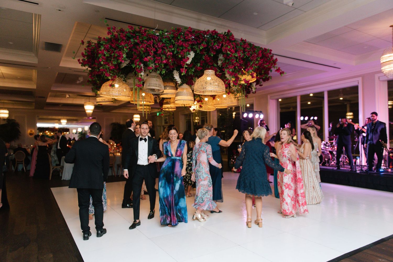 Wedding guests dance together on an illuminated dance floor under decorative floral arrangements at an elegant reception.