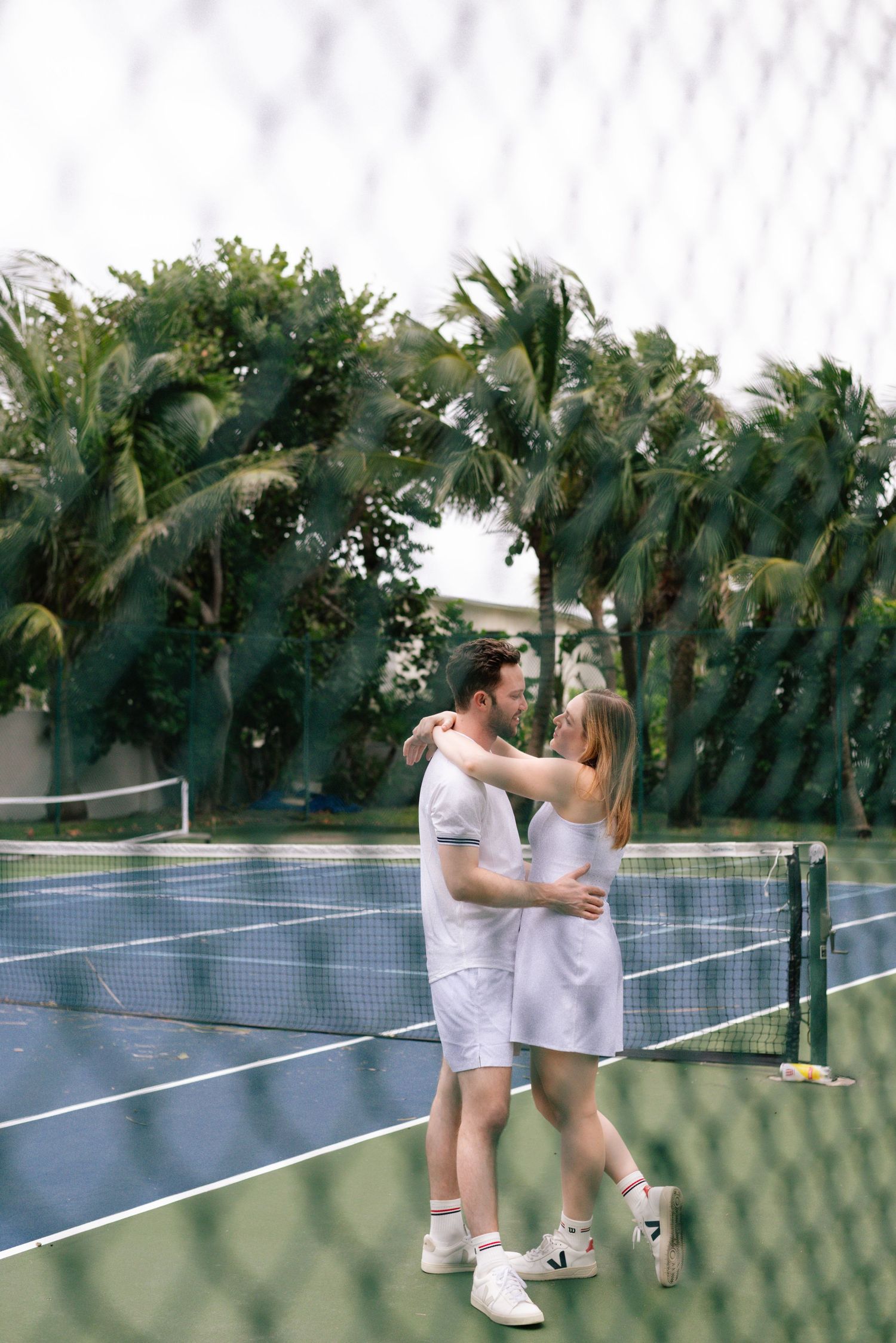 A couple in white tennis attire embraces near the net on a palm-lined tennis court.