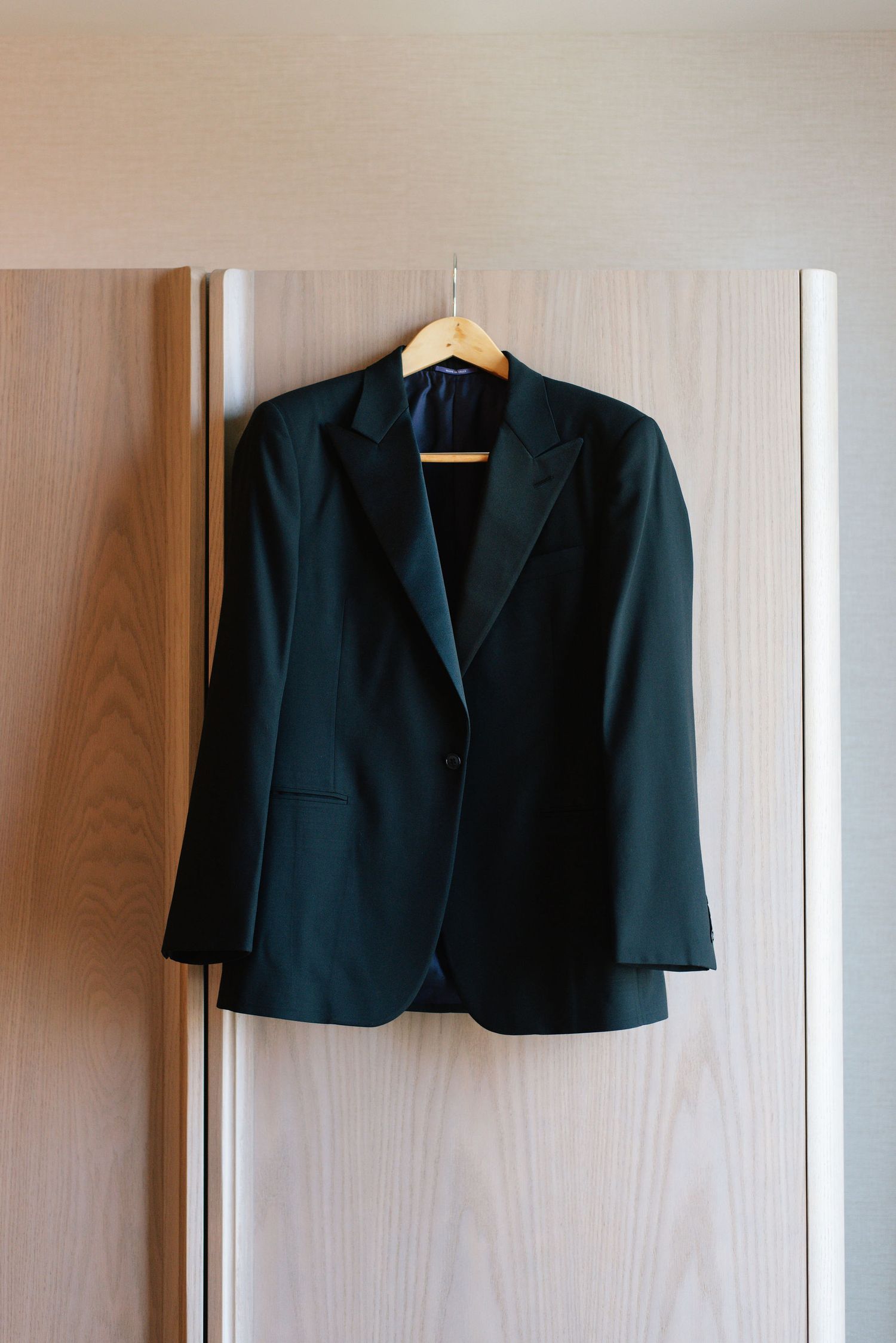 A black tailored blazer jacket hanging on a wooden wall against a light beige background.