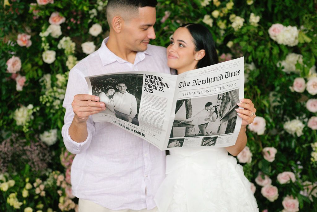 Couple in white outfits reading a newspaper together against a floral backdrop.