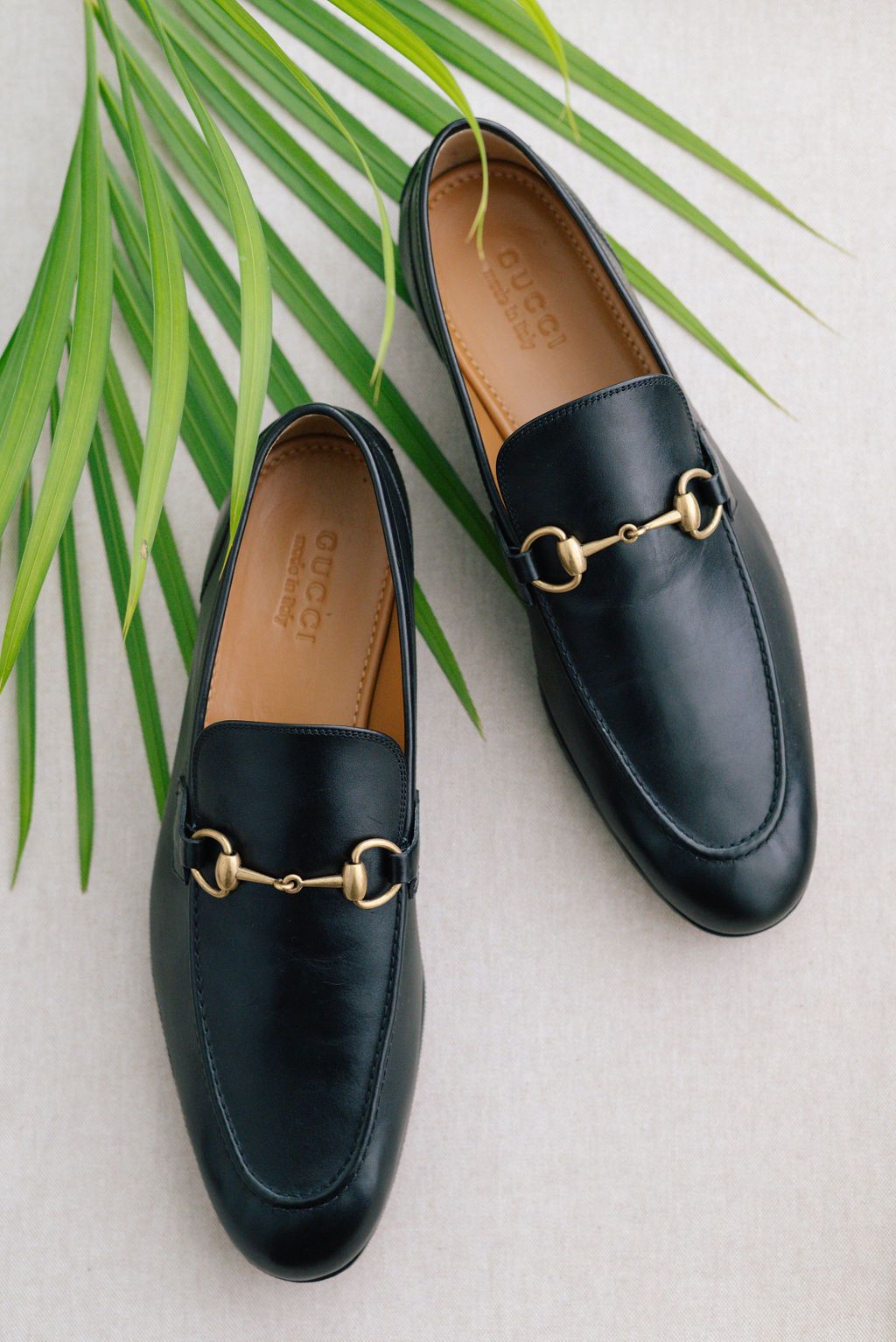 Black leather loafers with gold horsebit detail styled with tropical palm leaves.