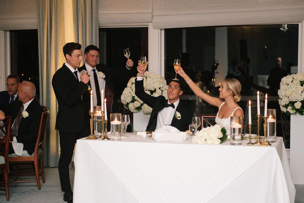Wedding guests raise their glasses in celebration at an elegant reception with candlelit tables.