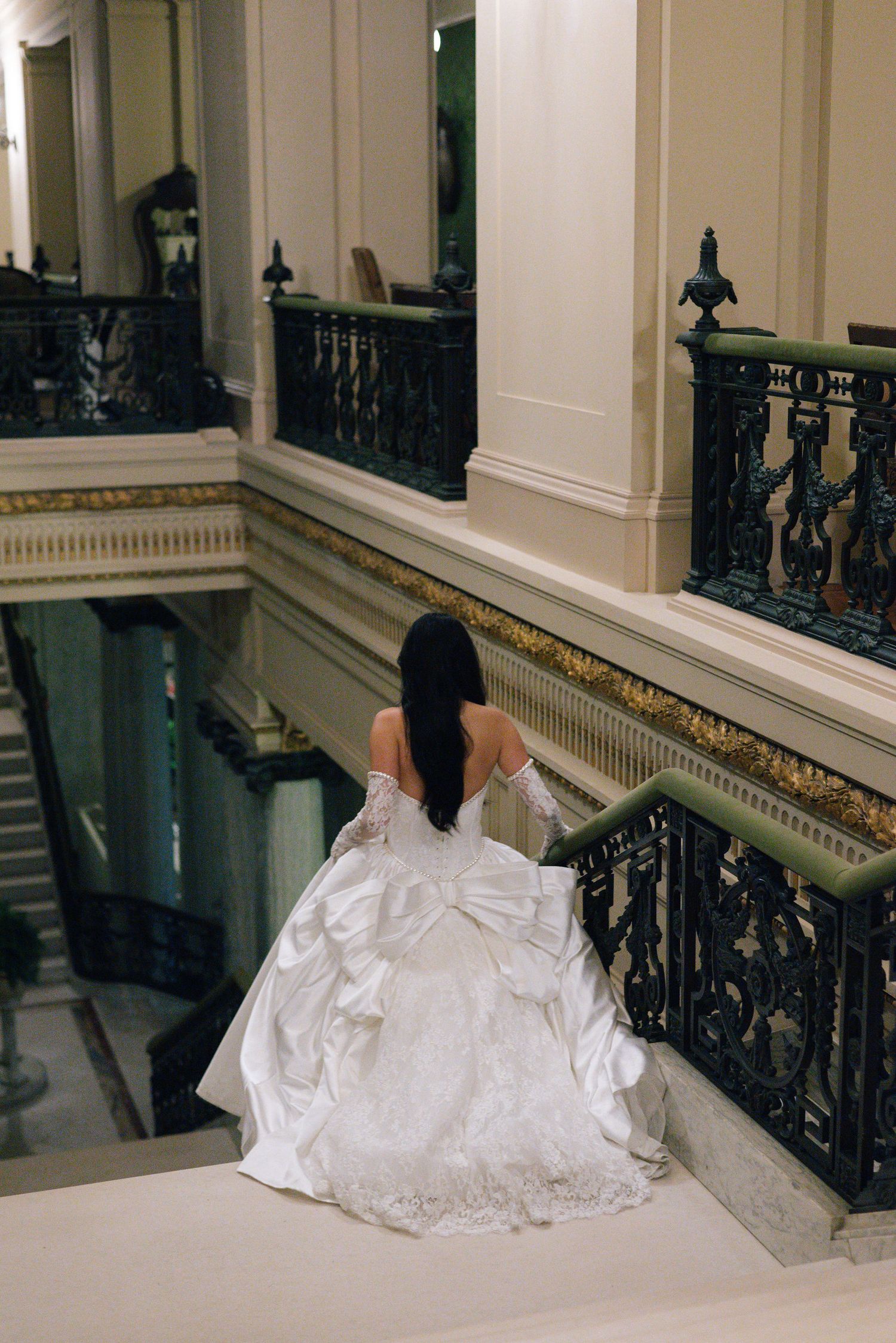 Bride in flowing white ballgown descending ornate staircase with detailed iron railings.