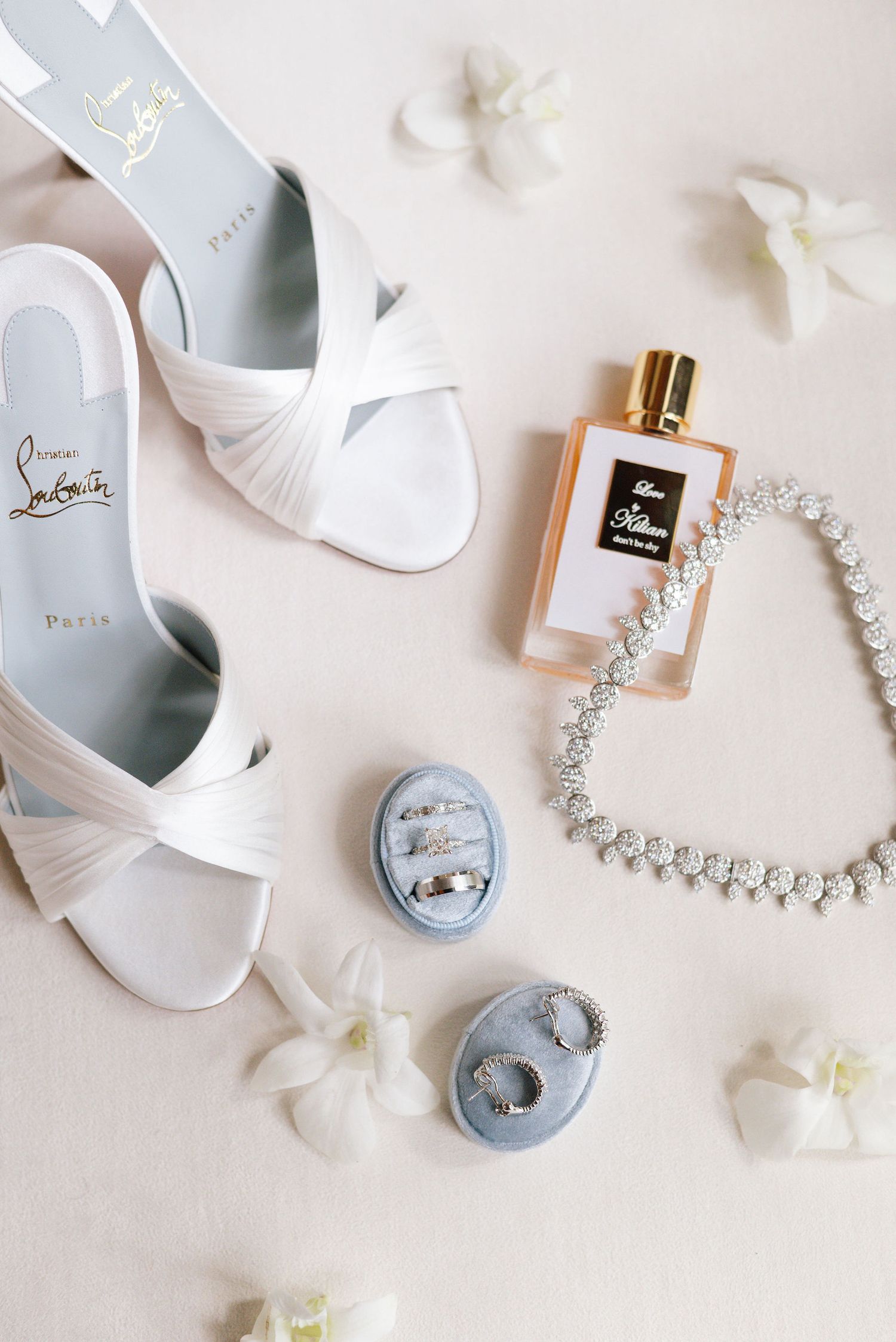 Bridal accessories including white sandals, perfume, crystal necklace and ring boxes with scattered petals.