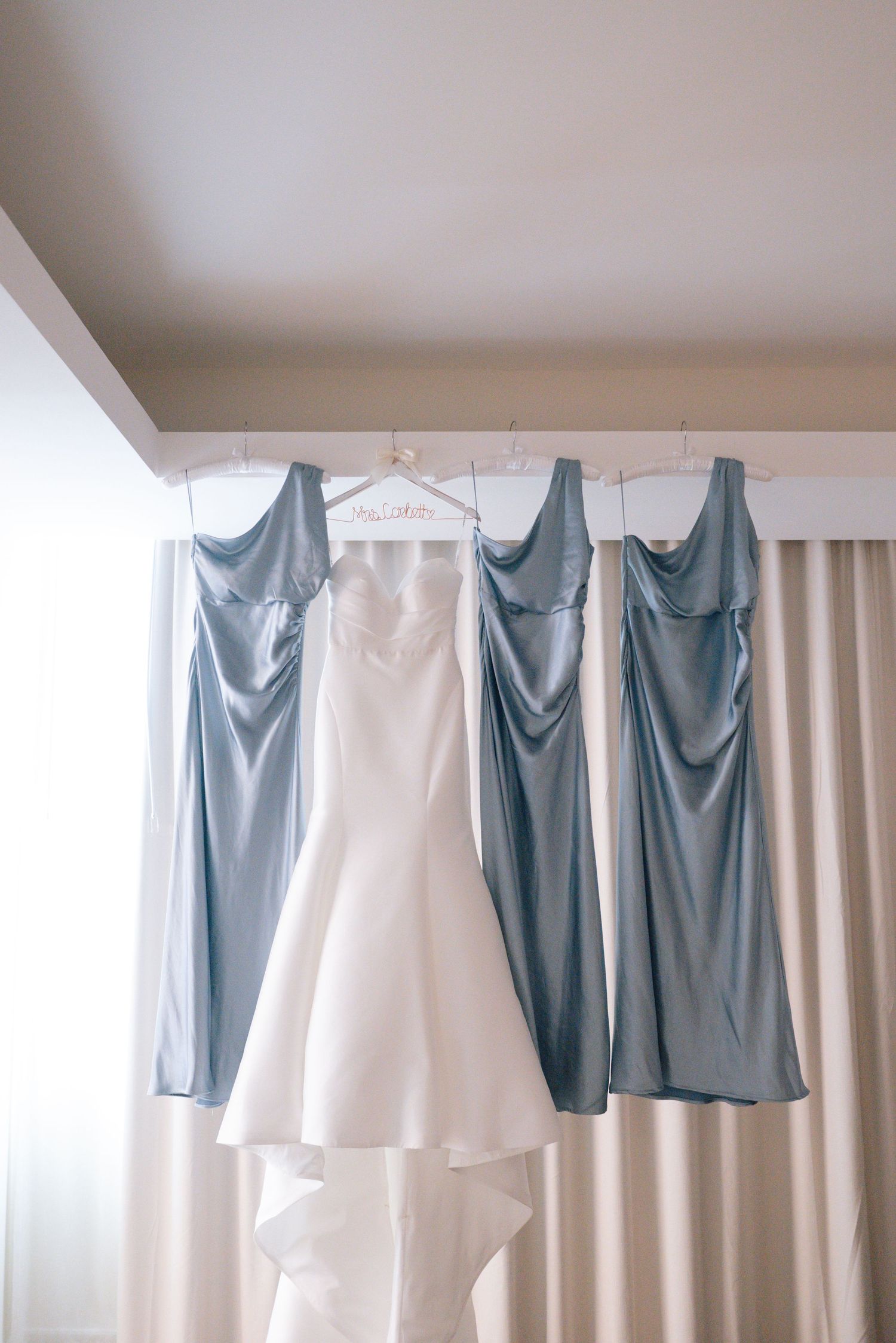 A white wedding dress and sage green bridesmaid dresses hanging on a curtain rod against a light wall.