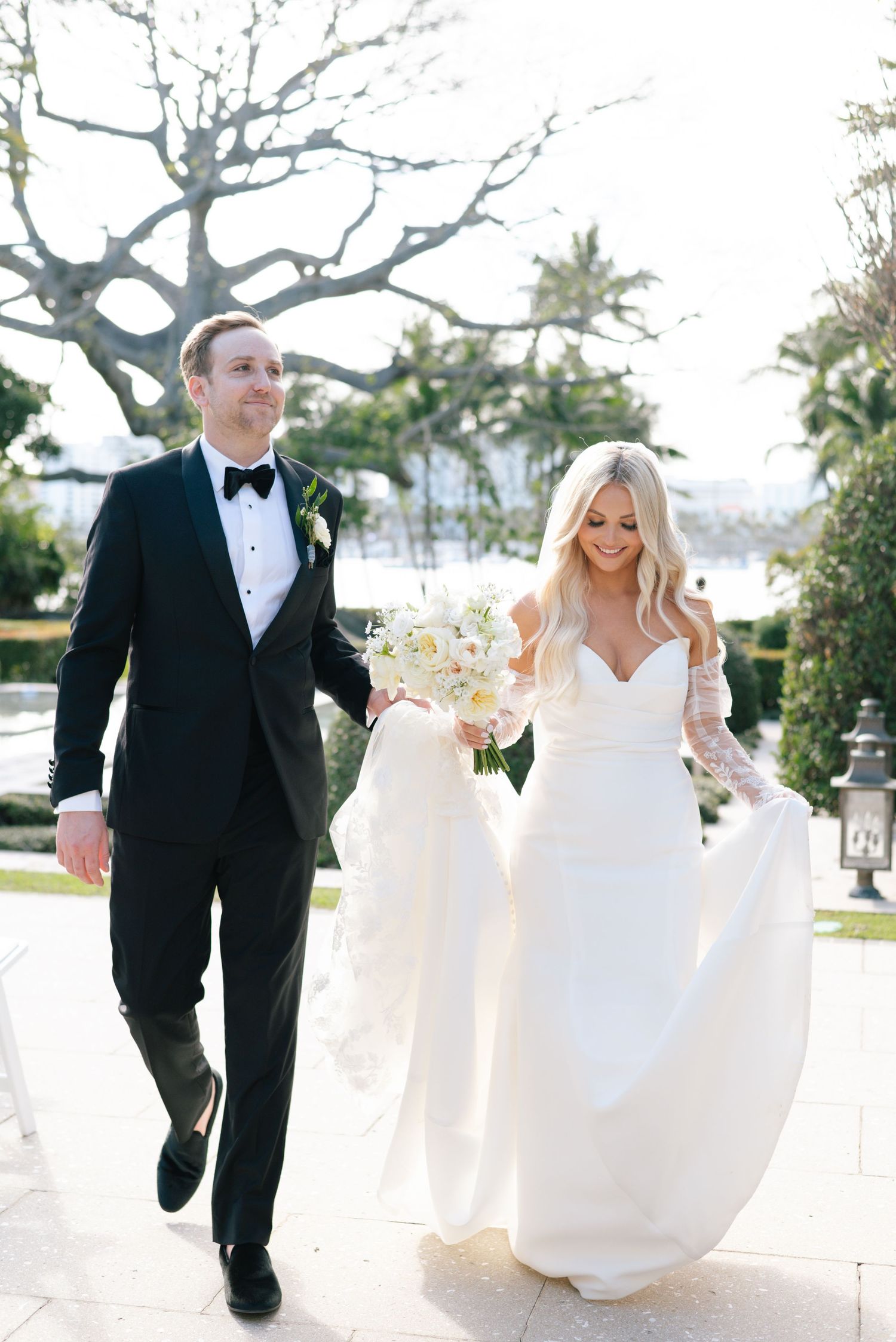 Wedding couple runs playfully through a sunlit garden path.