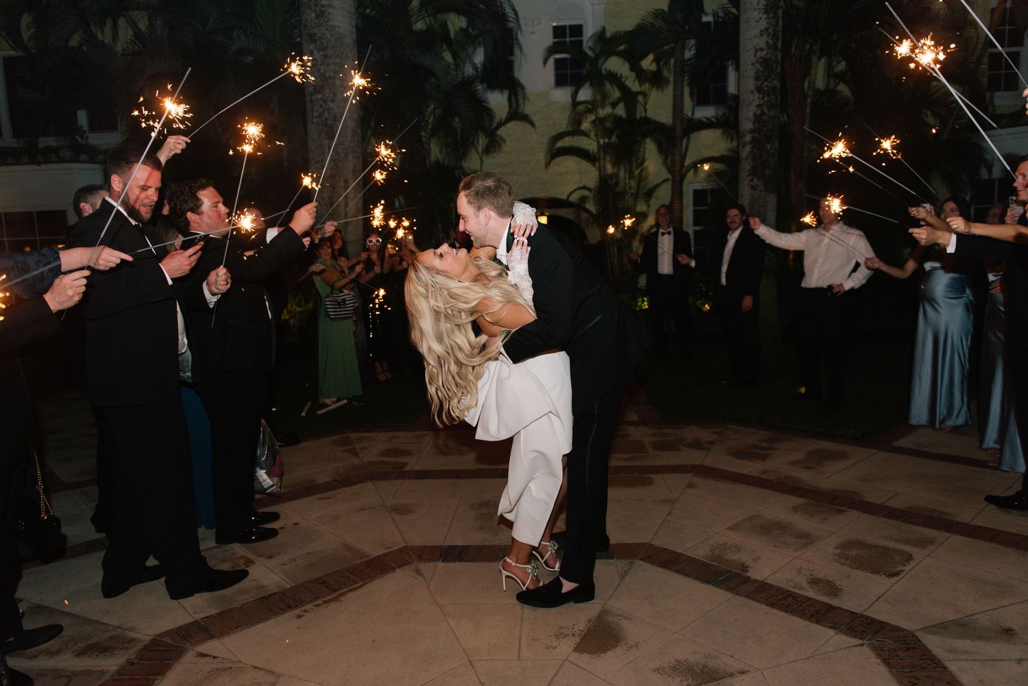 Wedding couple surrounded by guests holding sparklers during evening celebration.