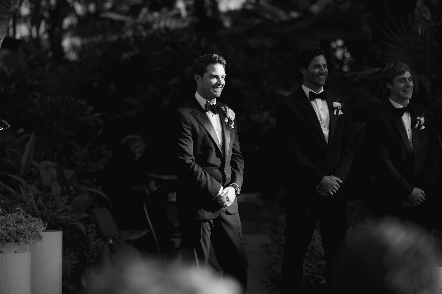 Three groomsmen in formal black and white tuxedos stand together during an elegant outdoor wedding ceremony.