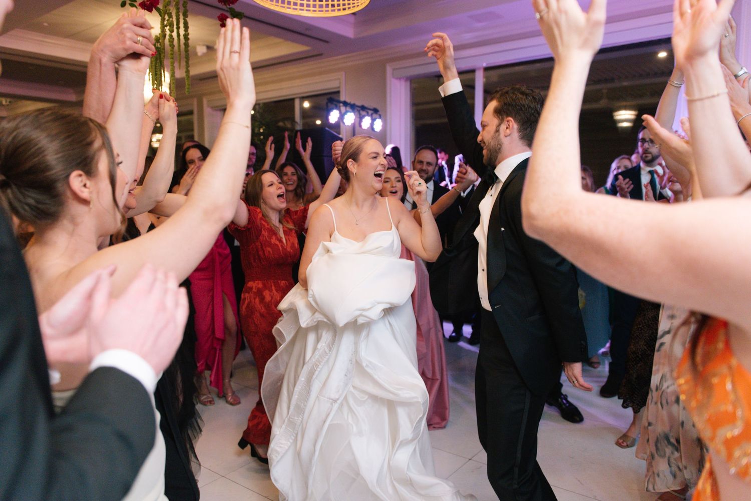 Wedding guests dance and celebrate with raised hands at a reception with purple lighting.