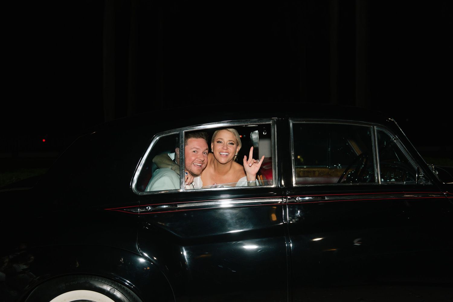 Happy newlyweds waving from vintage dark classic car during wedding night departure.