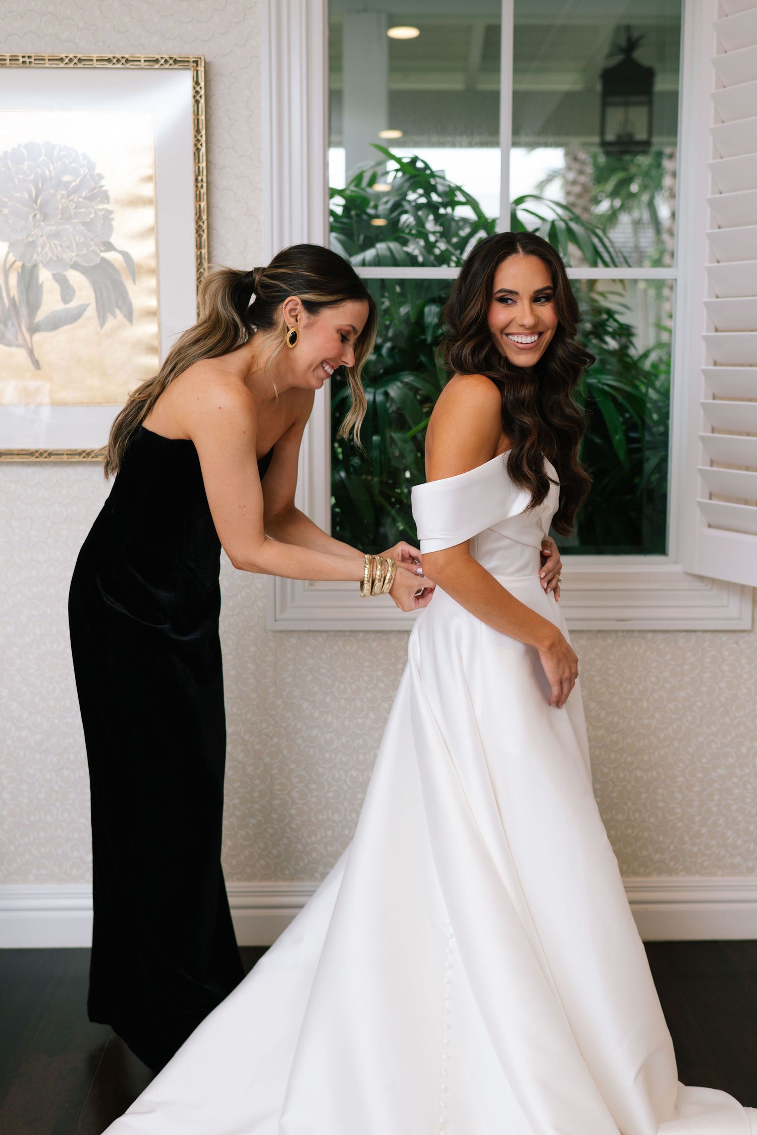 A bridesmaid in black helps adjust a stunning off-shoulder white wedding gown during pre-ceremony preparations.