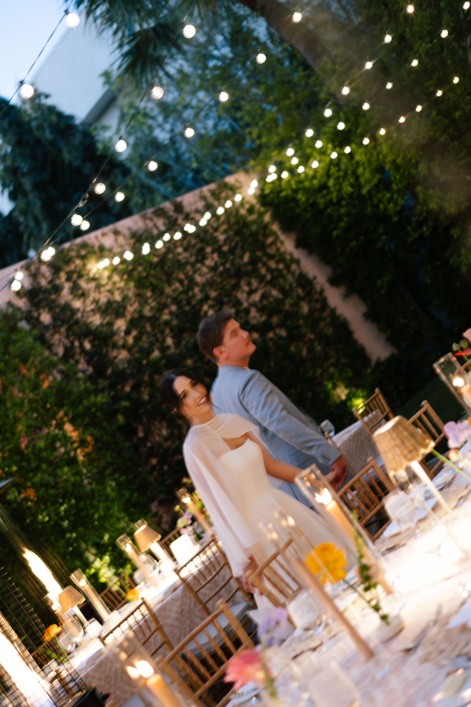 Garden reception tables set with white linens under twinkling string lights at dusk.