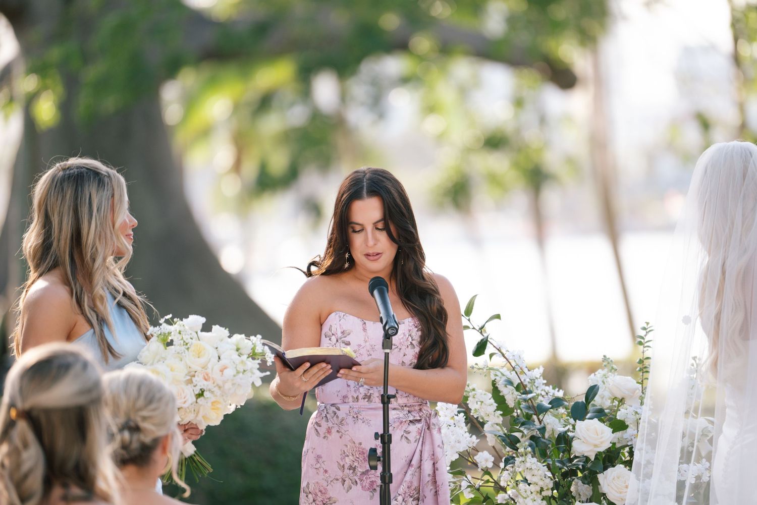 Wedding party member in a pink floral dress reads from a card during an outdoor ceremony surrounded by white flowers.