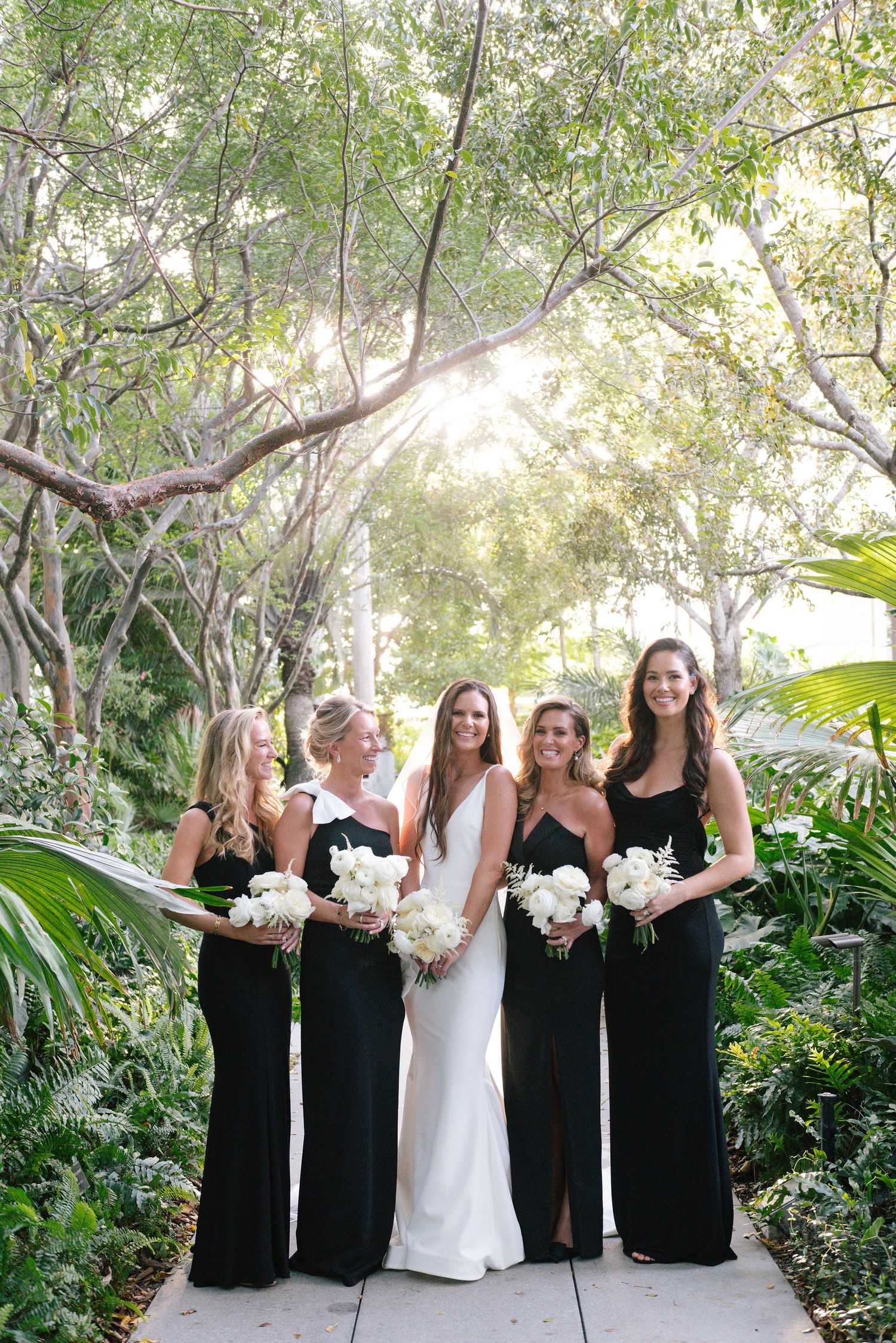 Bridal party in black dresses and white bouquets poses together in a lush garden setting.